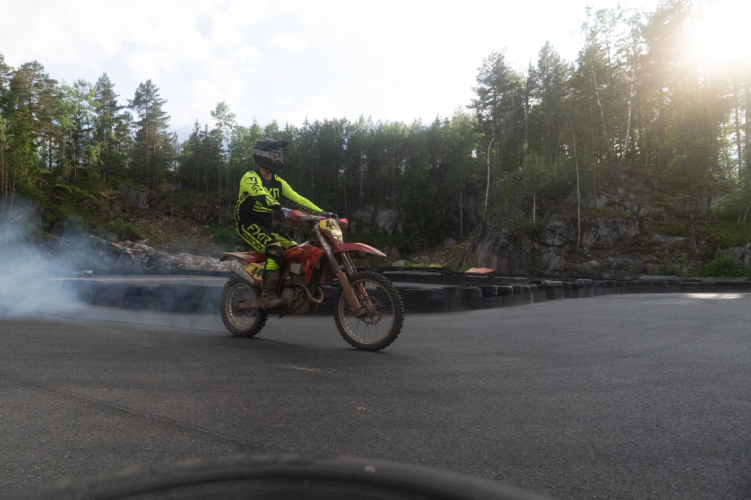 A motocross rider in bright yellow riding gear, wearing a black helmet, riding a red dirt bike on a paved racetrack with trees and rocks in the background, and smoke behind the bike.