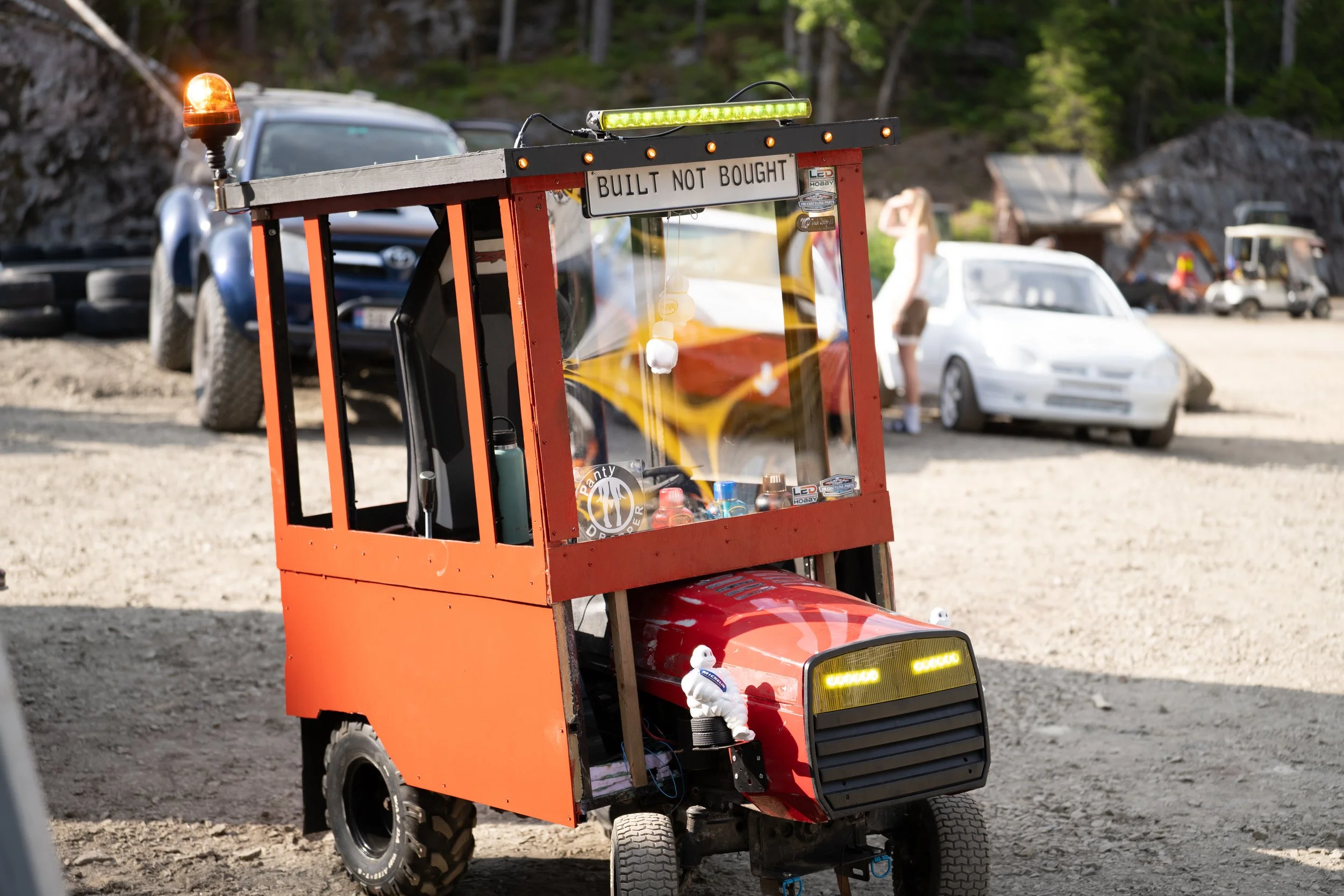 A small red vehicle with a sign that reads "Built Not Bought," resembles a miniature car or go-kart, in a dirt lot with people and cars in the background.