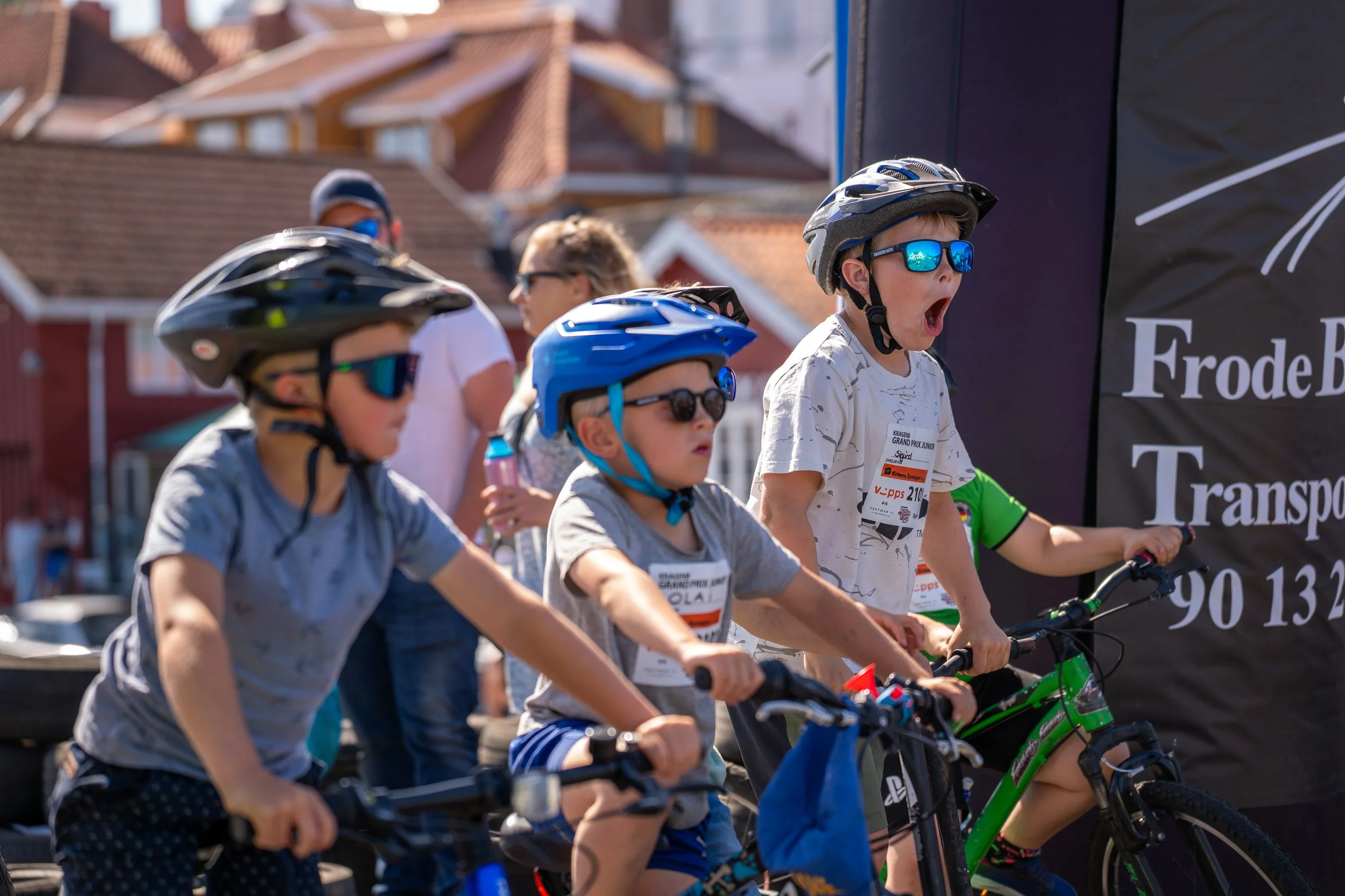 Young boys wearing helmets and sunglasses riding bicycles at a race event, with spectators and a black banner in the background.