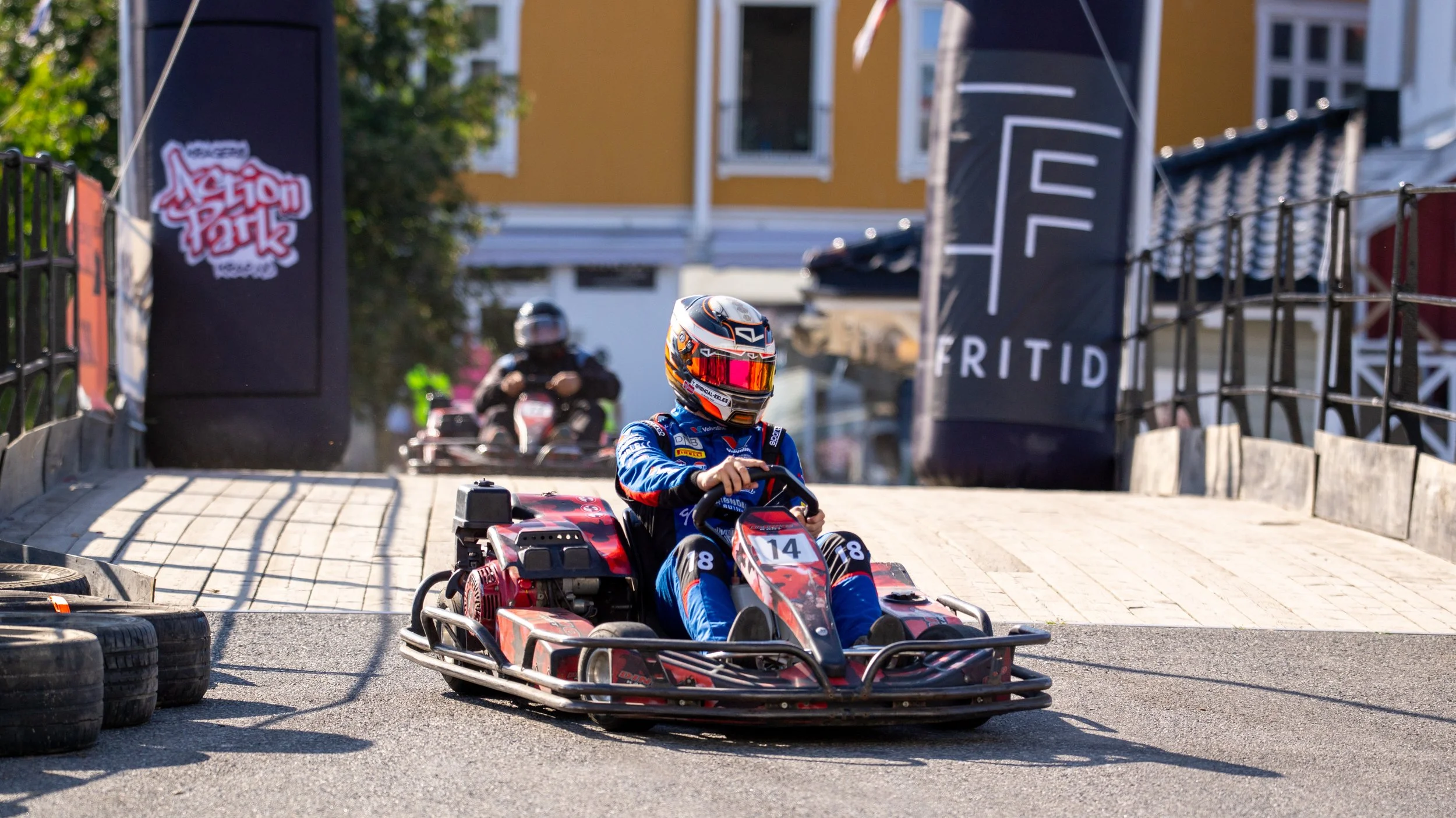 A person driving a red and black go-kart on an outdoor track, wearing a blue racing suit and a helmet with a red visor, with another person in a go-kart in the background.