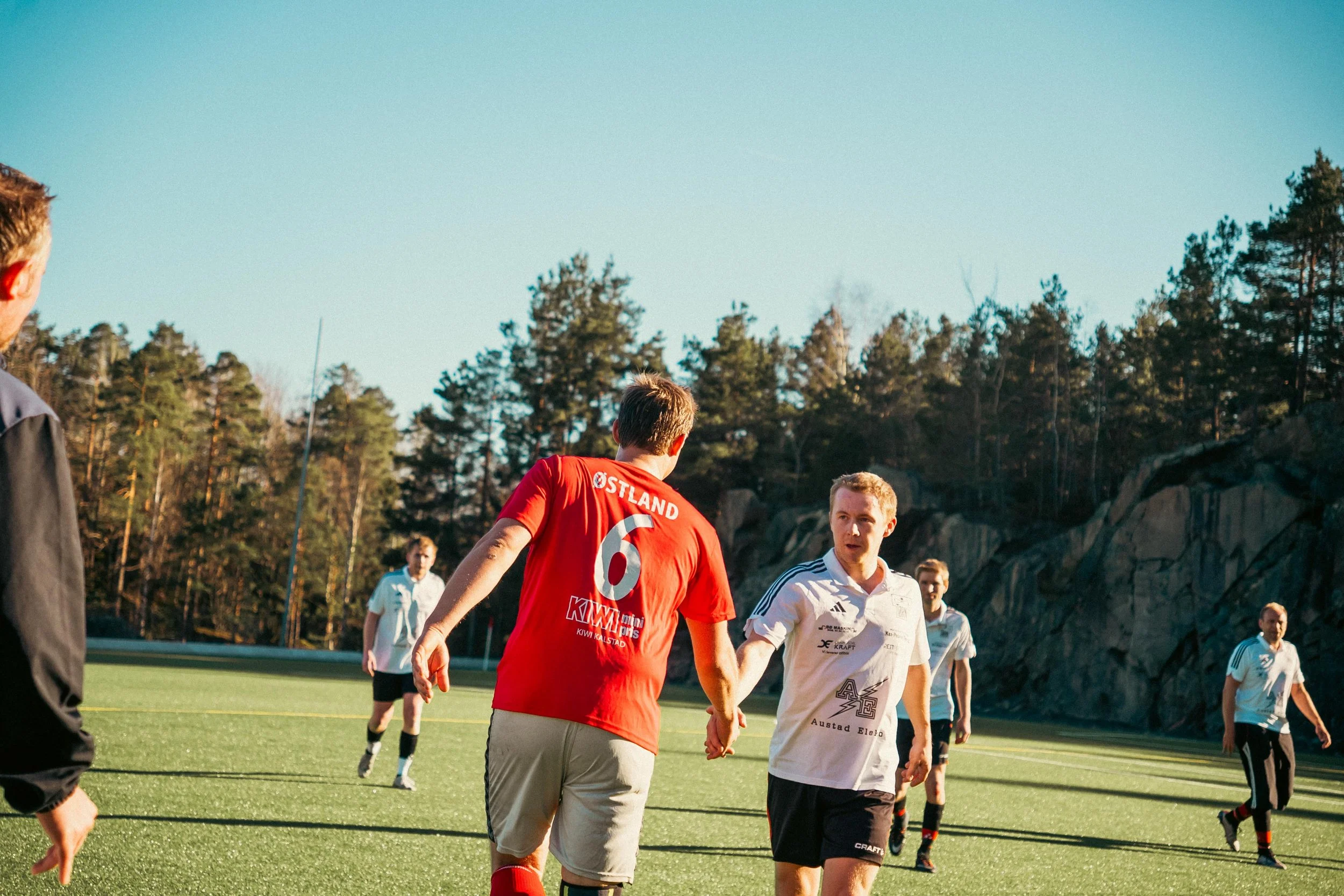 A group of soccer players on the field, with two players in foreground shaking hands, during a game on a sunny day with trees in the background.