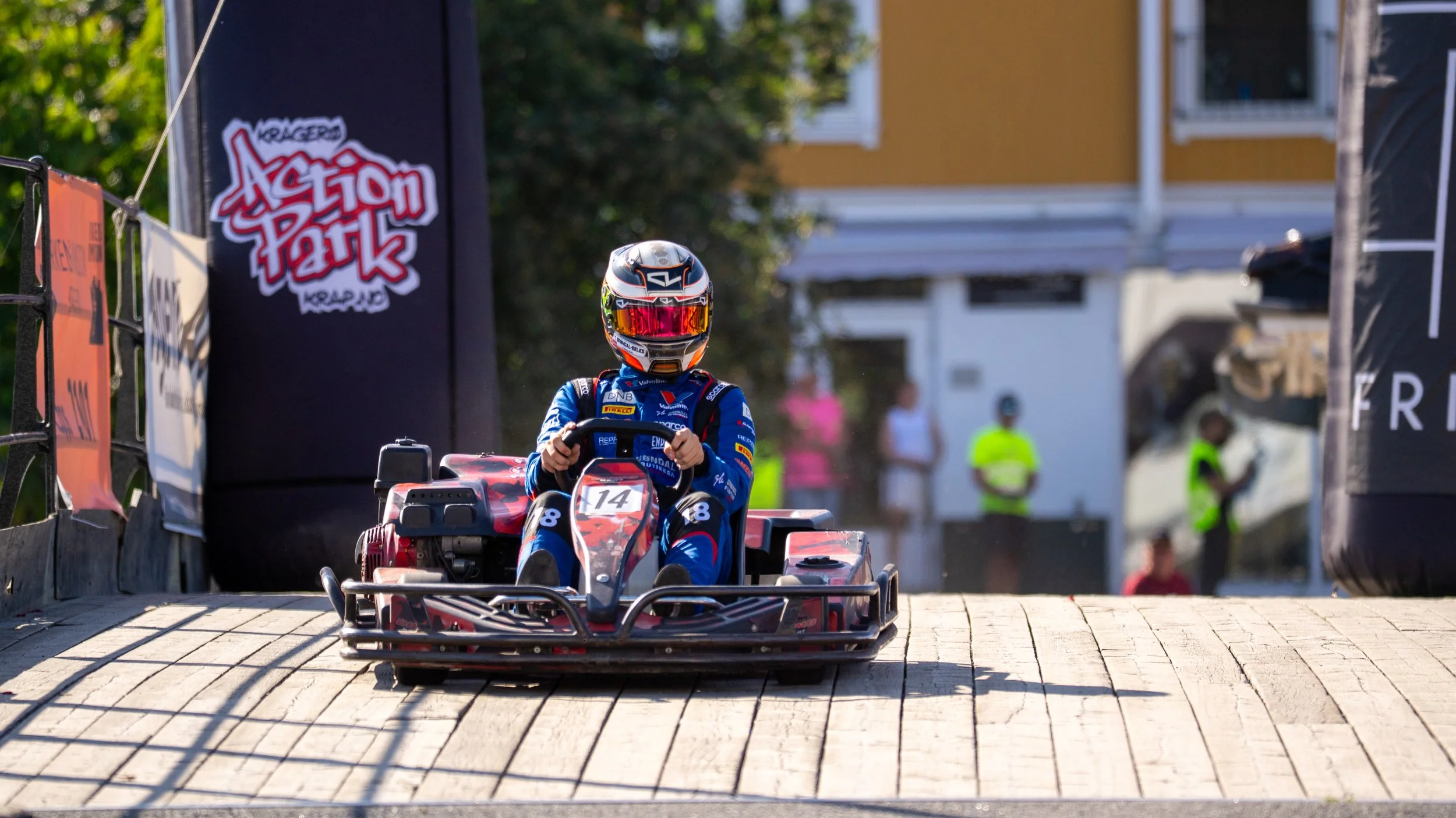 A person wearing a blue racing suit and helmet driving a go-kart on a wooden track at Action Park, with a yellow building and people in the background.