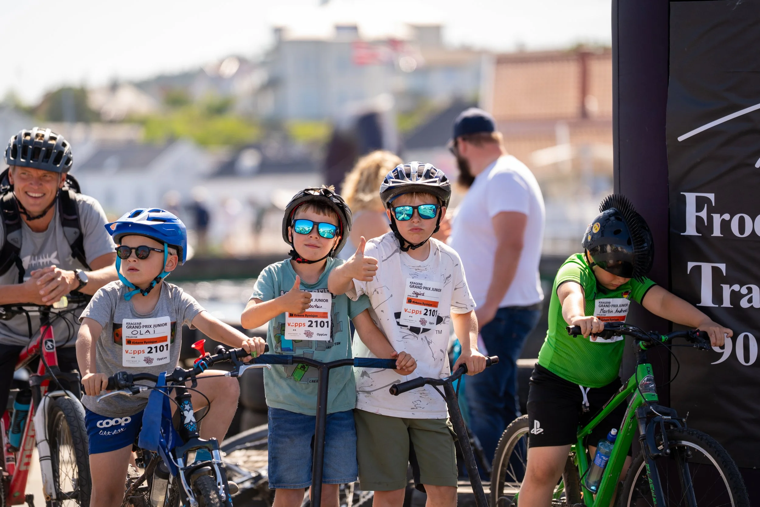 Children with helmets and sunglasses on bikes, giving thumbs up at a racing event near the water.