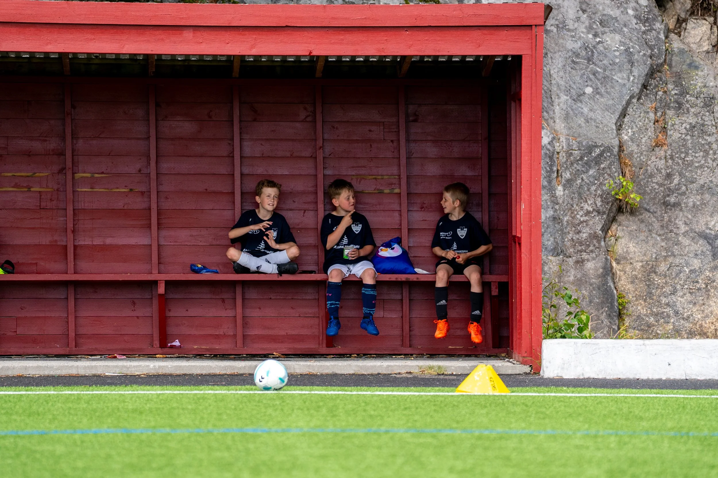 Three young boys sitting on a red wooden bench in a soccer dugout, watching a soccer game on a green field with a ball and yellow training cone in the foreground.