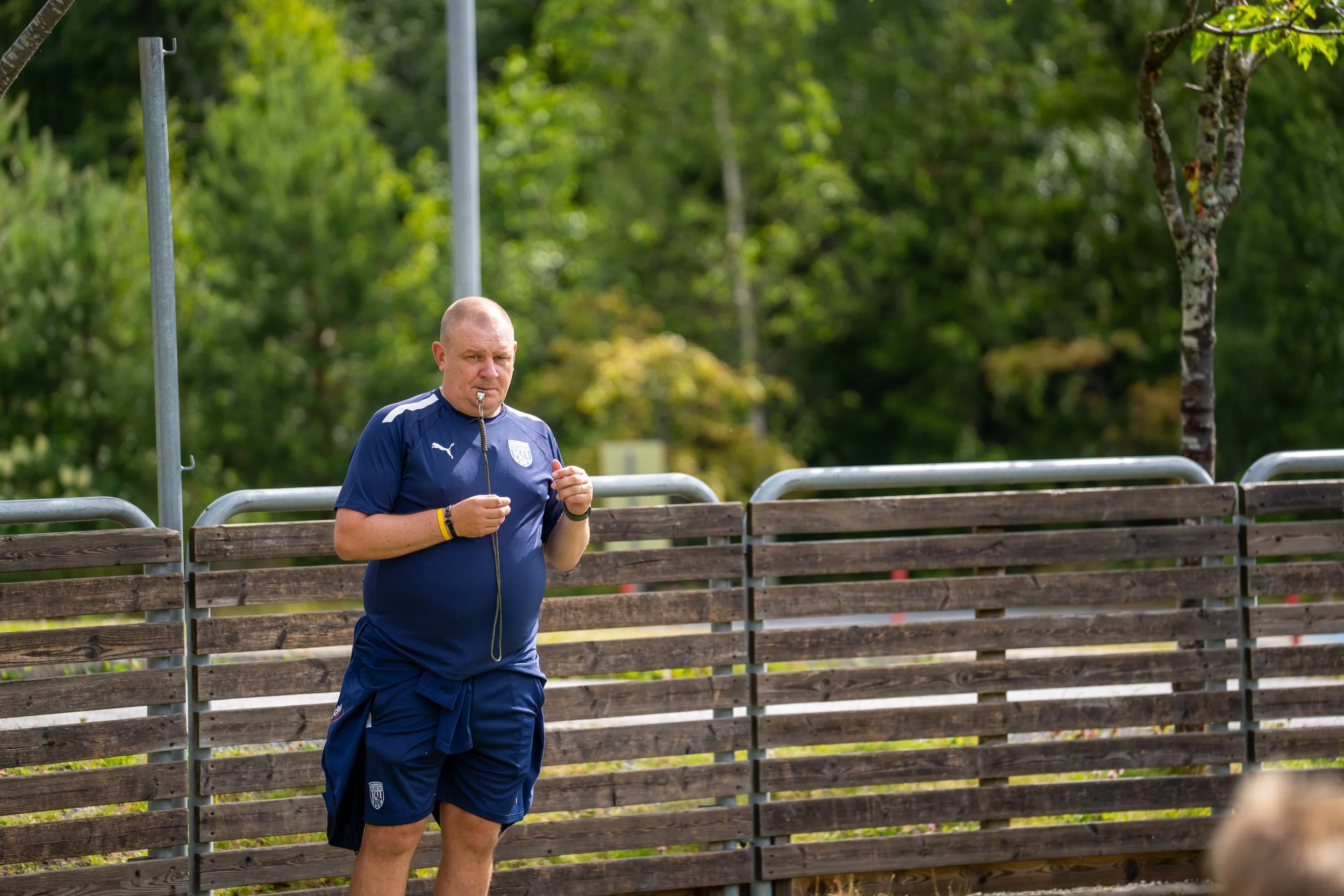 A man dressed in a blue sports uniform with a whistle around his neck, standing outdoors next to a wooden fence, with green trees in the background.