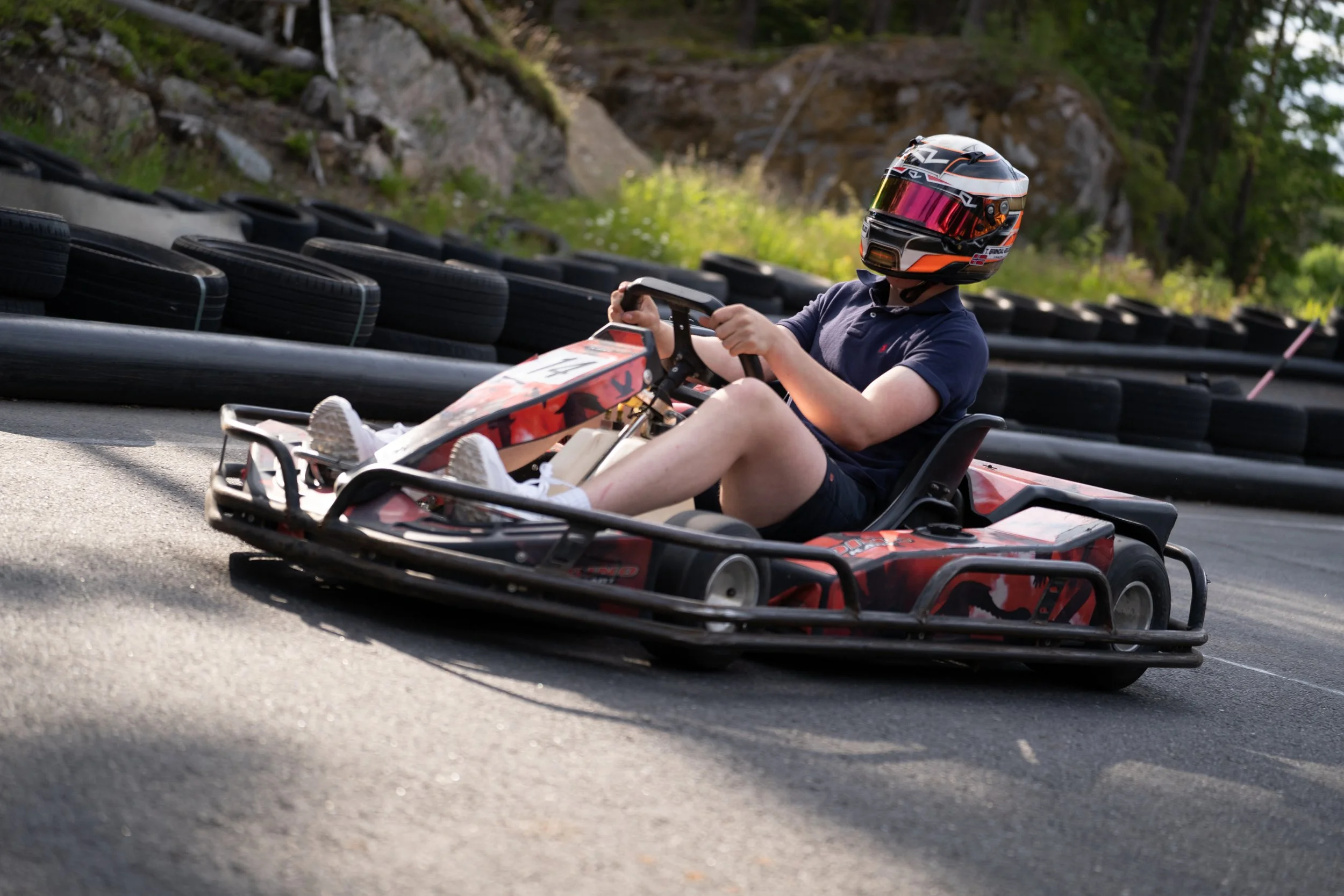 A person wearing a helmet and navy shirt driving a go-kart on an outdoor race track, with tire barriers on the side and trees in the background.