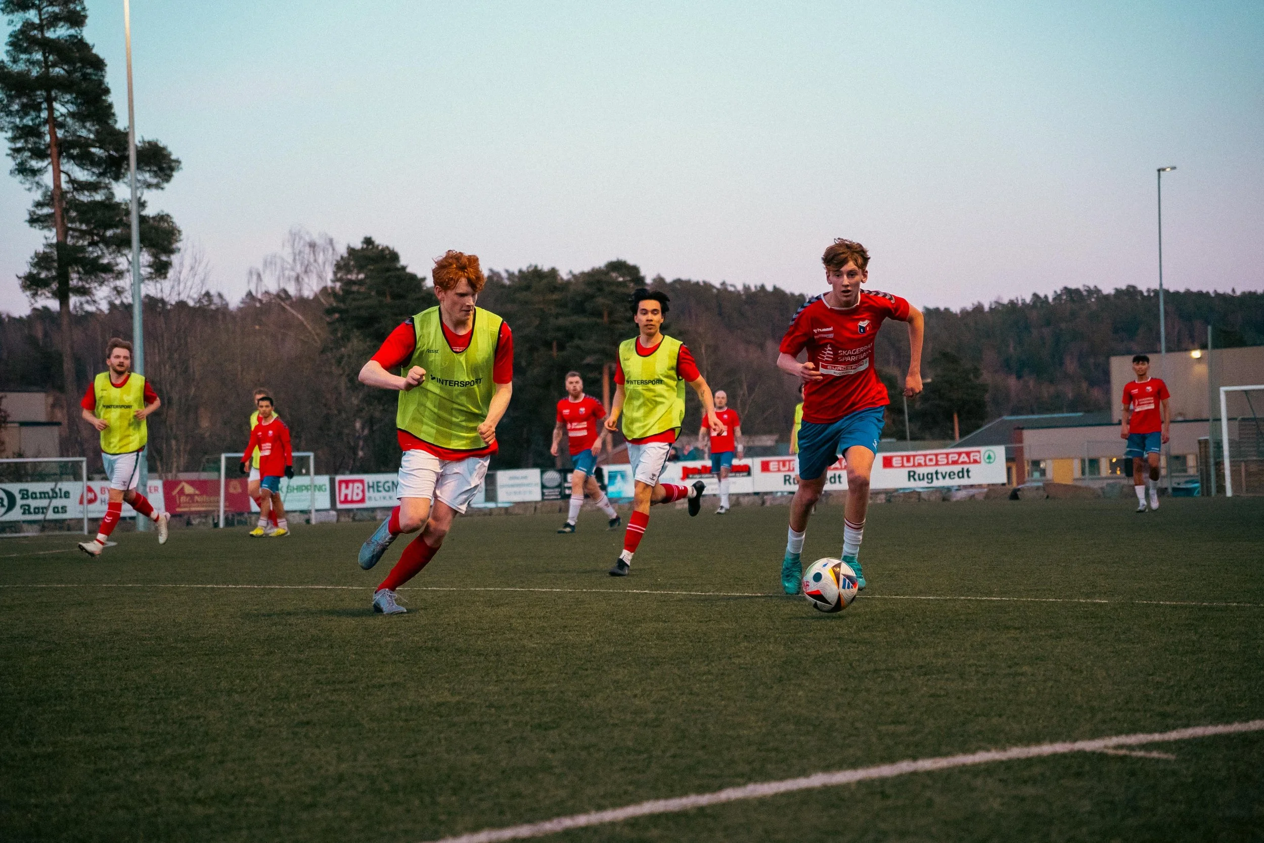 Soccer players practicing on a field during dusk, with some wearing yellow vests and others in red jerseys, with trees and advertisements in the background.
