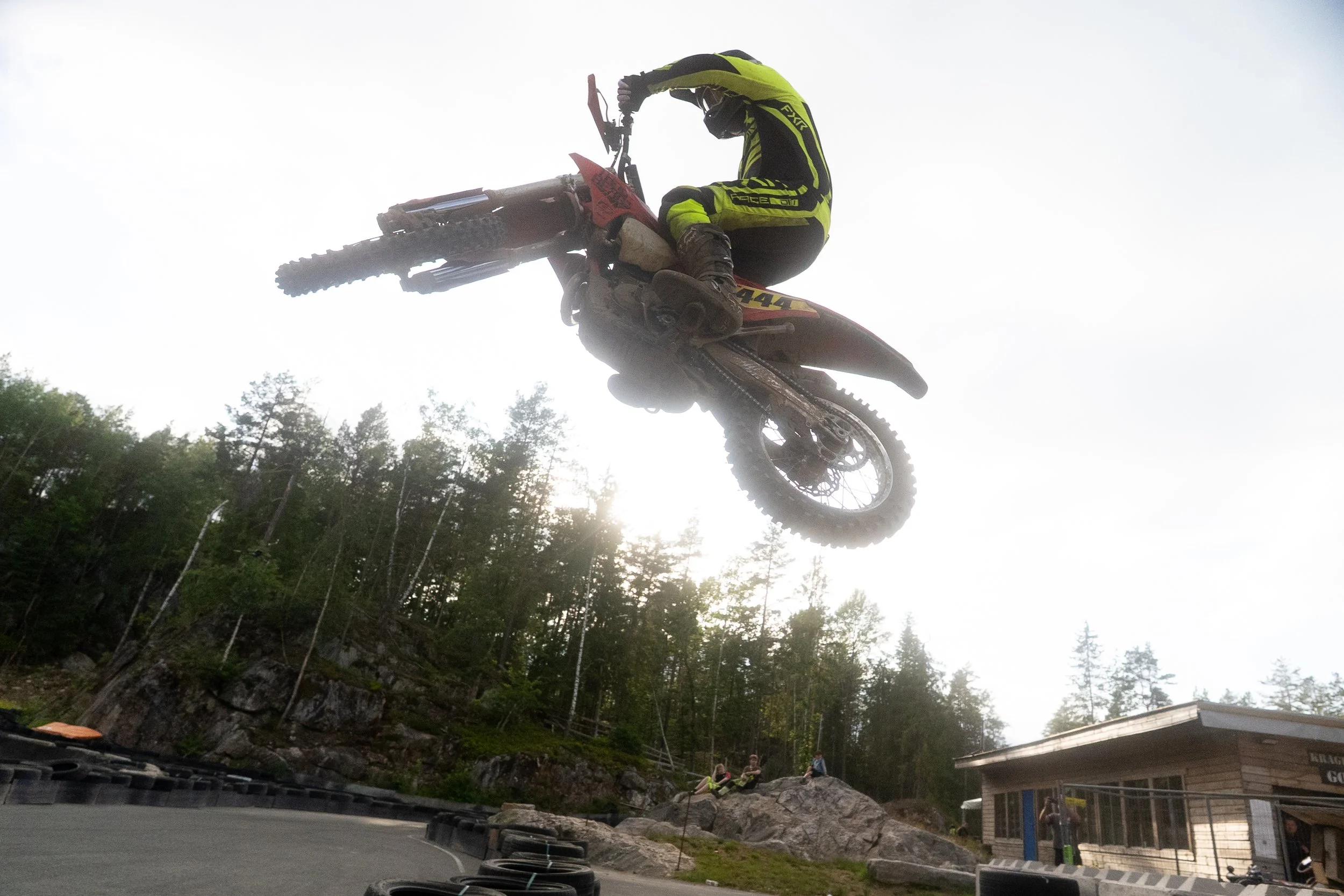 Motocross rider in yellow and black gear jumping a dirt bike in midair over a mountain track, with trees and a building in the background.