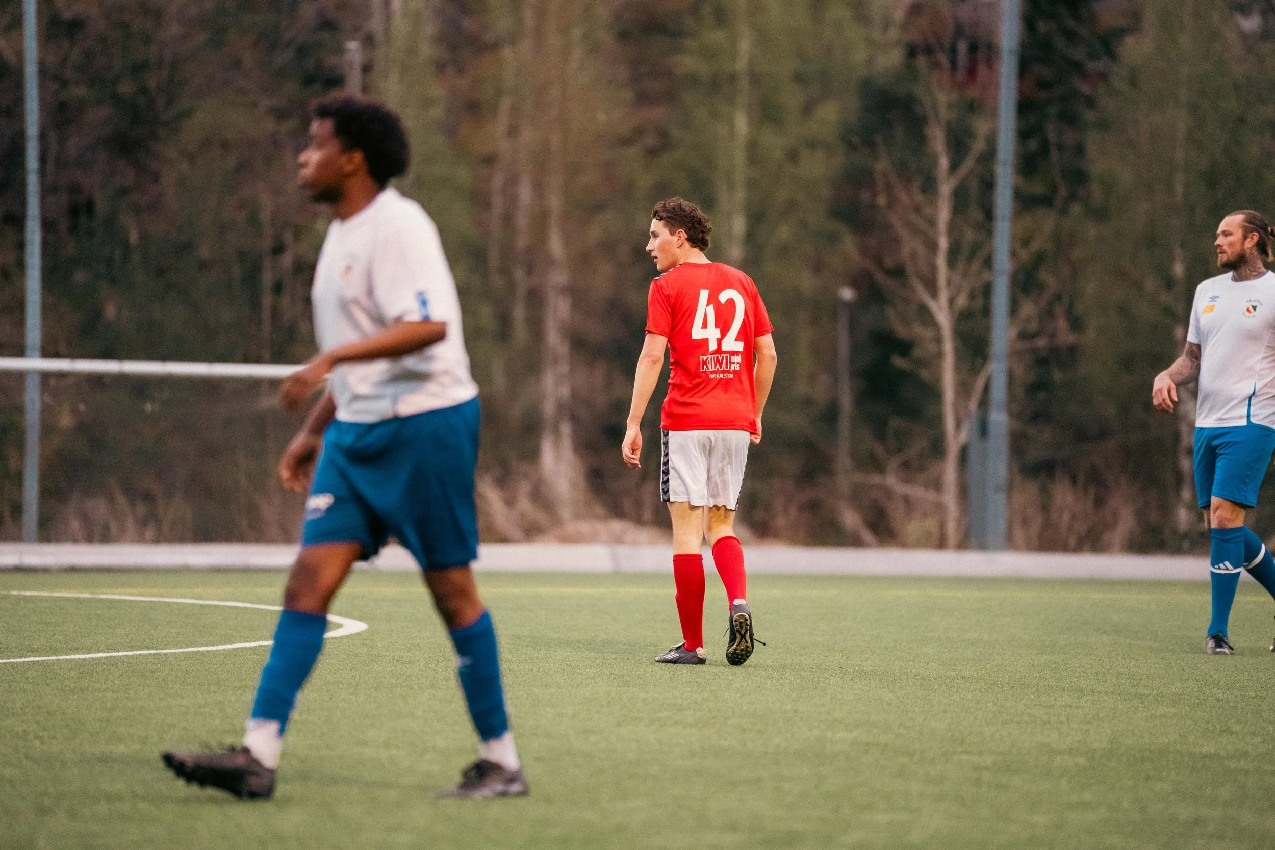 Three soccer players on a field, one near the center wearing a red jersey with the number 42, and the other two in white and blue uniforms, one in the foreground and one in the background.