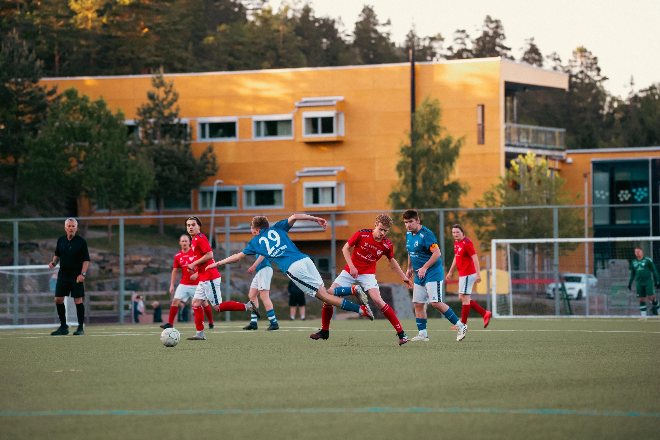 Soccer players in blue and red uniforms compete on a green field during a match at sunset, with a modern yellow building and trees in the background.