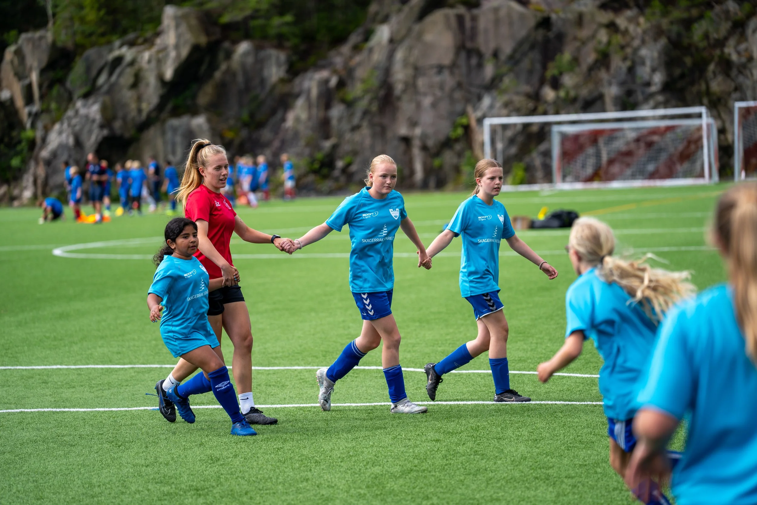 Young girls holding hands on a soccer field during a game, with one girl in a red jersey and others in blue, surrounded by a grassy field and goalposts, and a rocky hillside background.