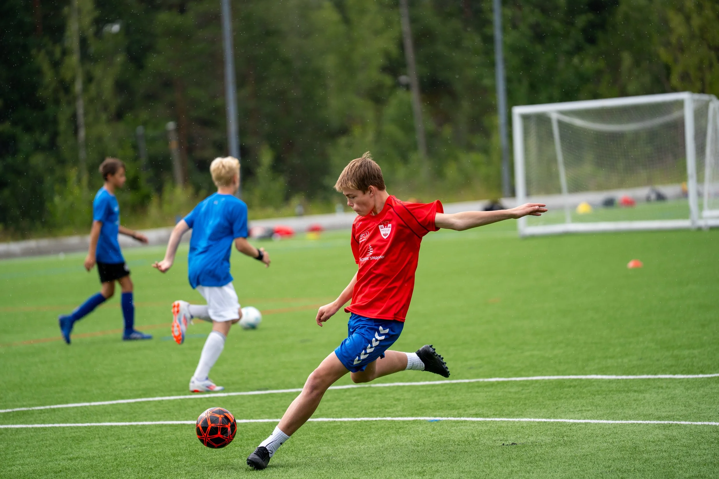 Young boys playing soccer on a field, with one boy in a red jersey kicking a black and red soccer ball while two others in blue jerseys run in the background, near goalposts and with trees behind.