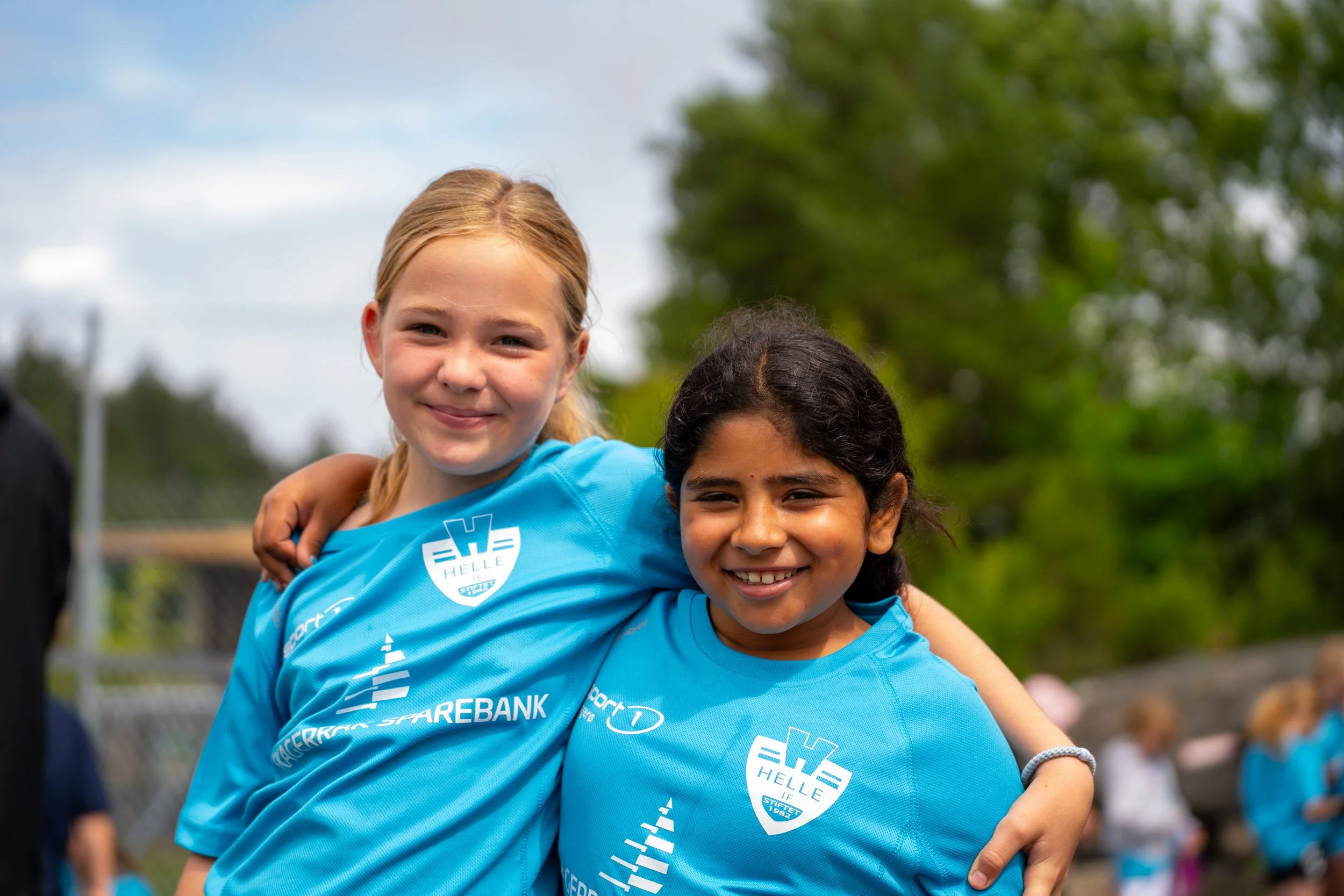 Two young girls in blue sports jerseys with arm around each other smiling outdoors on a cloudy day.