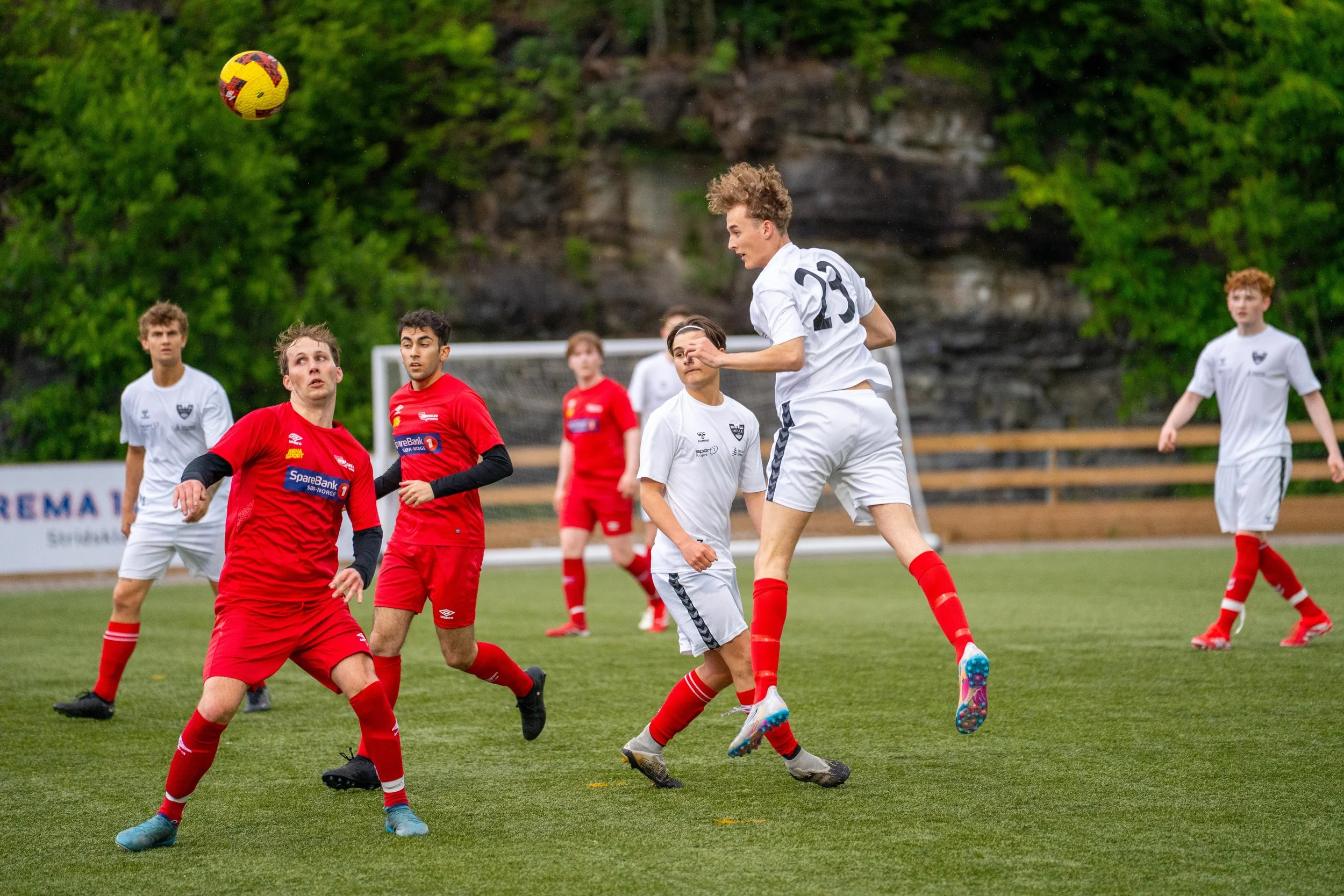 Soccer match with players in red and white uniforms on a grassy field, focusing on a player in white jumping to head the ball, surrounded by other players watching.