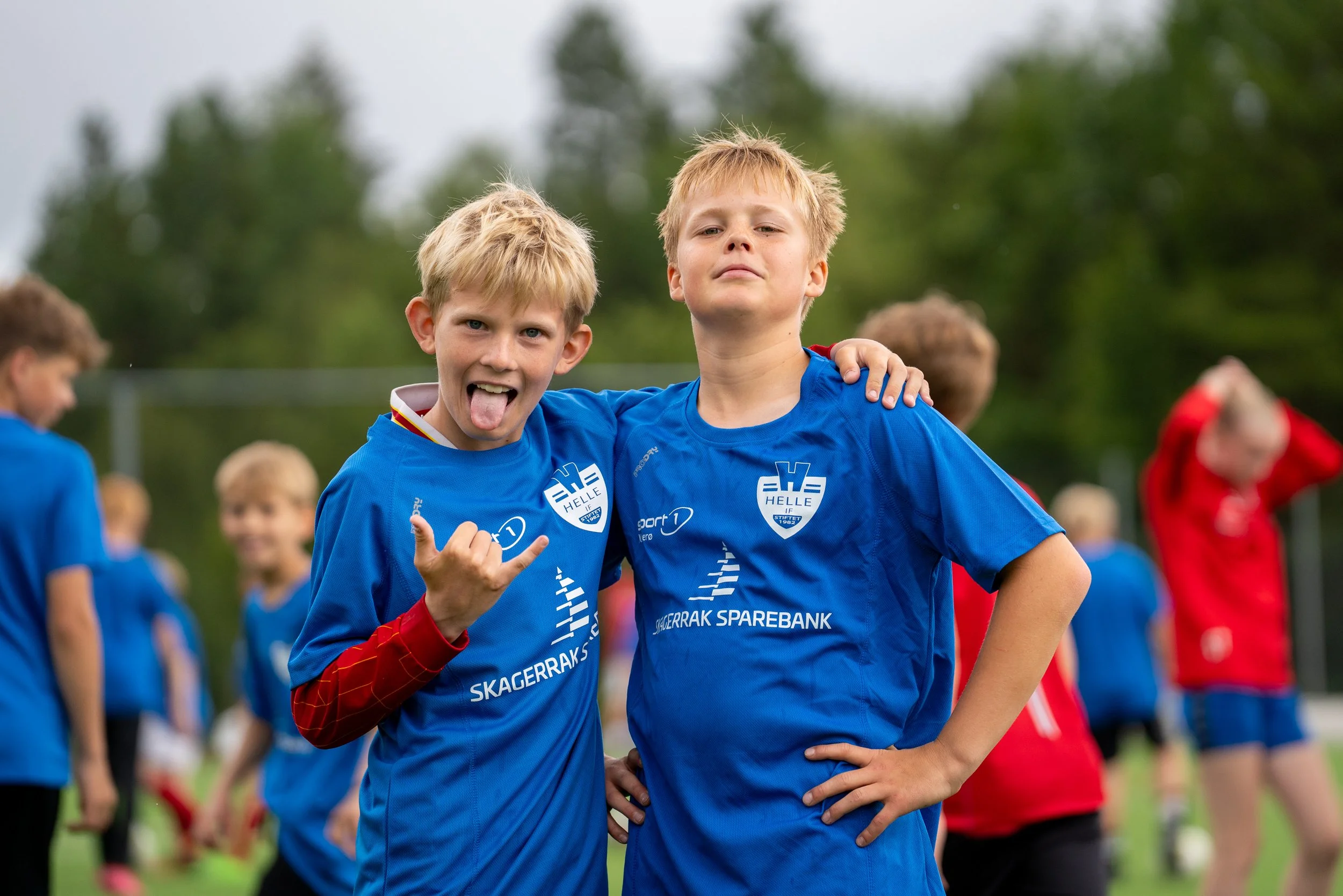Two young boys in blue sports jerseys posing for a photo on a soccer field, with trees in the background. One boy is sticking his tongue out and making a hand gesture, while the other has his hand on his hip and looks confident.