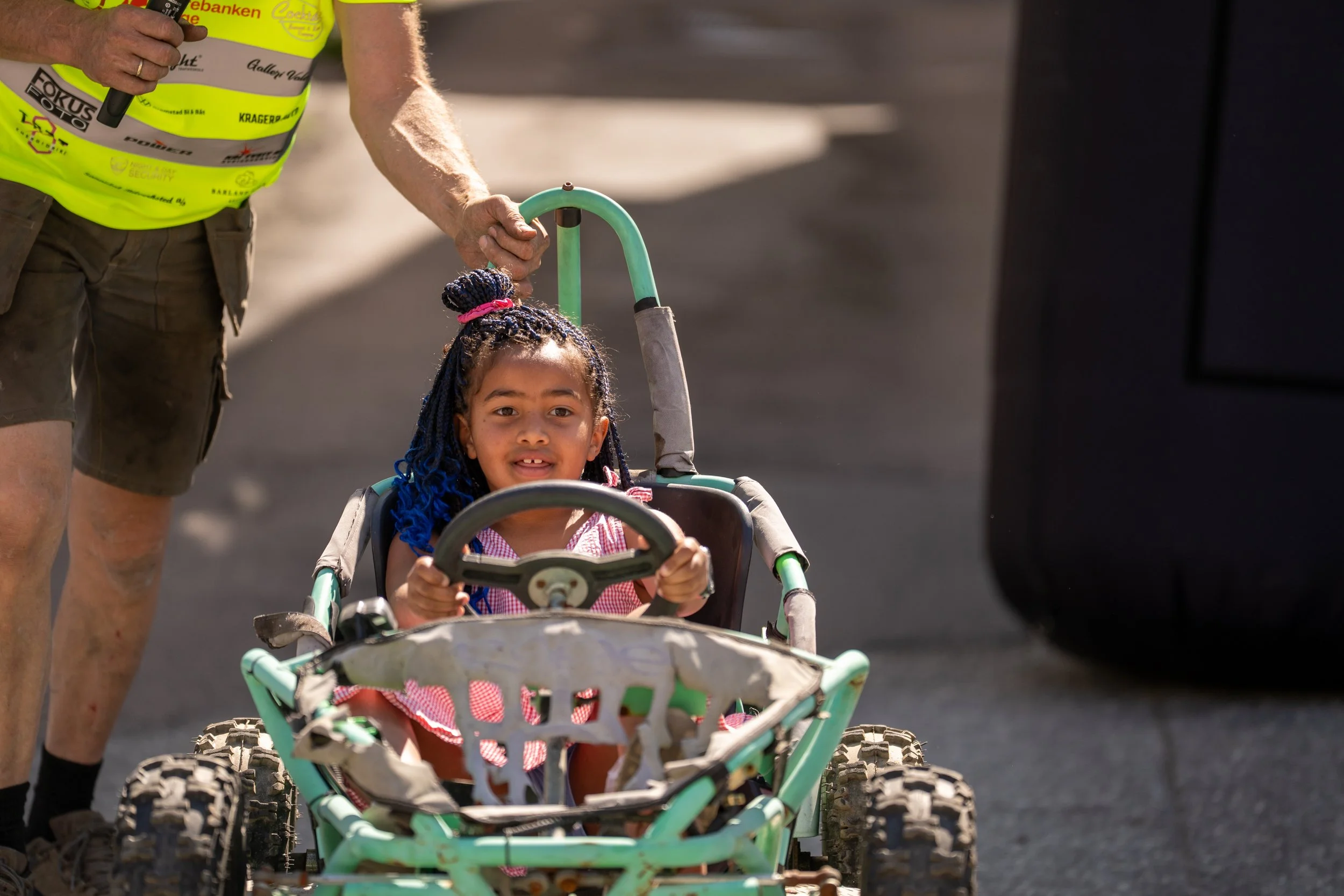 A young girl with braided hair and colorful beads sitting in a green pedal go-kart at an outdoor event, holding the steering wheel, with an adult man standing beside her holding onto the handlebar for support.