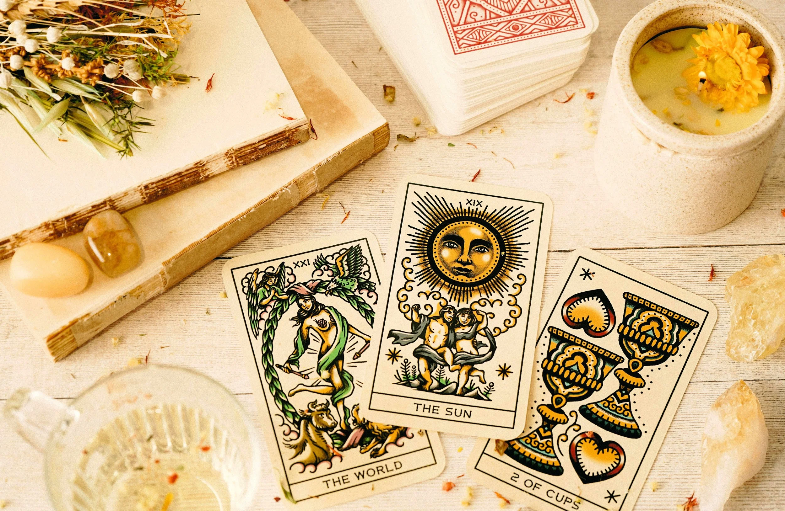 Tarots, bougies, pierres, et fleurs séchées sur une surface en bois blanc, peut-être pour une séance de lecture de tarot ou de méditation.