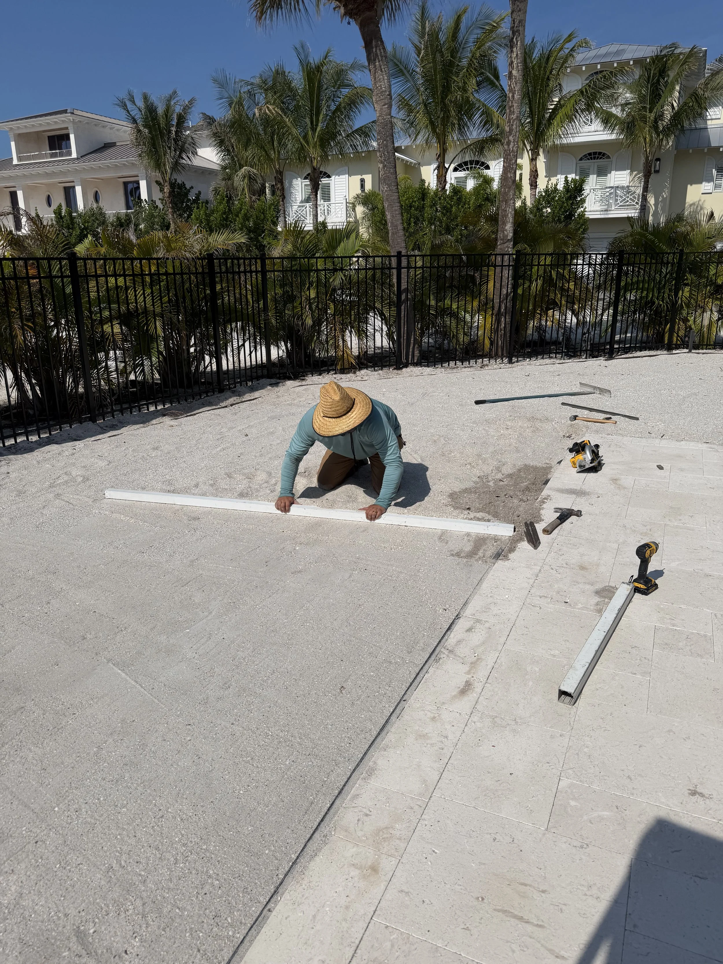 A person wearing a straw hat and a blue long-sleeve shirt kneels on a sandy surface, working with a long white metal piece. Tools and construction materials like a saw, hammer, drills, and other hardware are scattered around. In the background, there