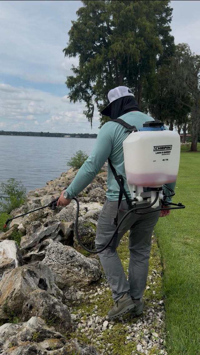 A person wearing a gray cap, a face covering, and a backpack sprayer is spraying liquid along a rocky shoreline near a body of water, with trees and a cloudy sky in the background.