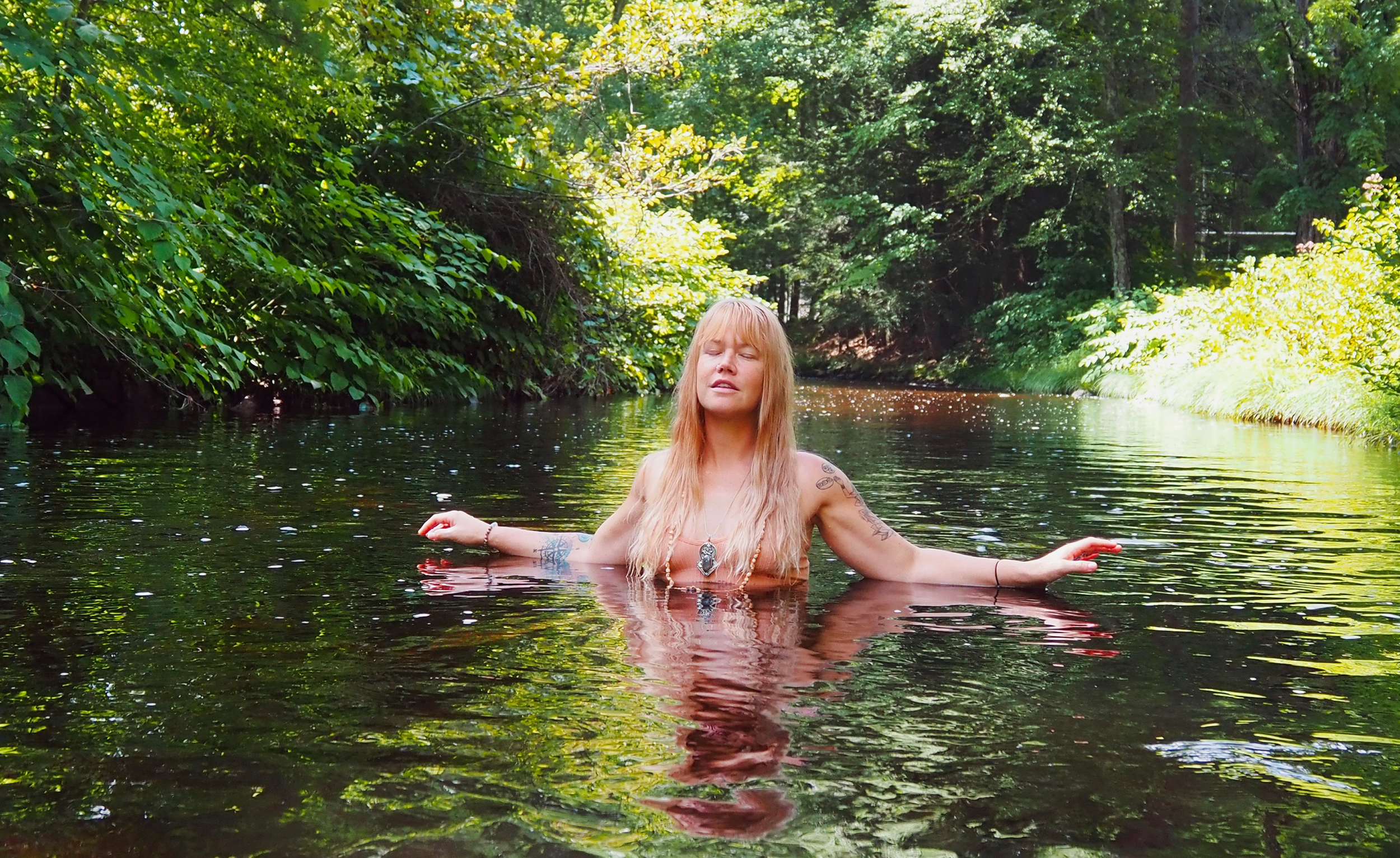 A woman with long blonde hair and tattoos is standing waist-deep in a river surrounded by lush green trees and foliage, with sunlight filtering through the leaves.
