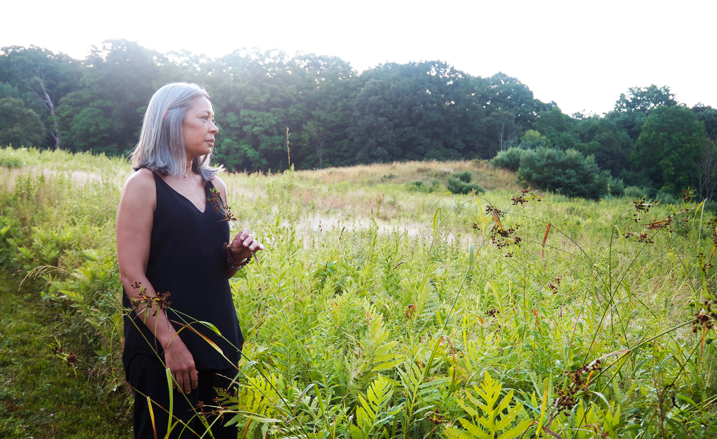 An older woman with gray hair wearing a black sleeveless top standing in a lush green field with trees in the background.