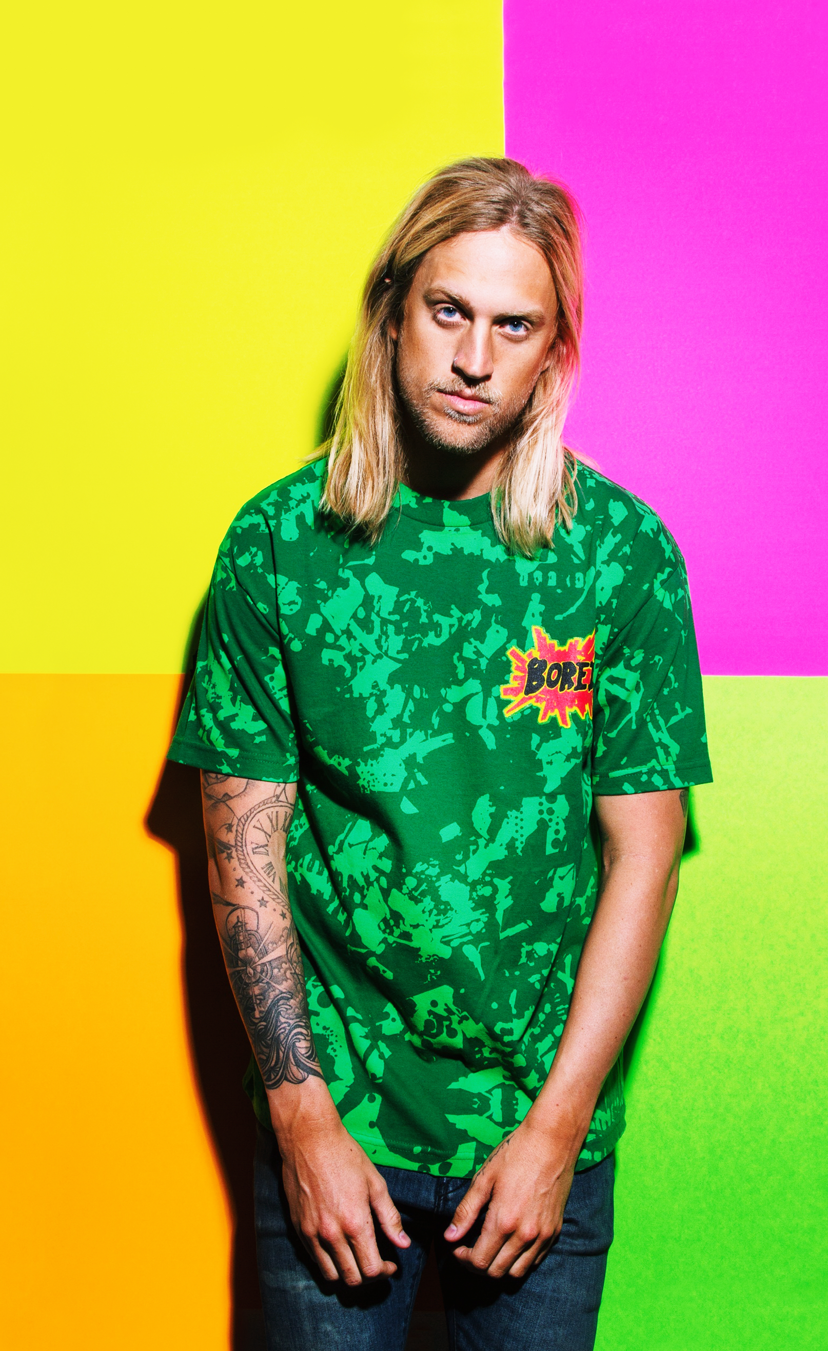 A man with long blond hair, wearing a green patterned T-shirt with a comic book style "BORE" graphic, standing against a colorful background with yellow, pink, and green sections.
