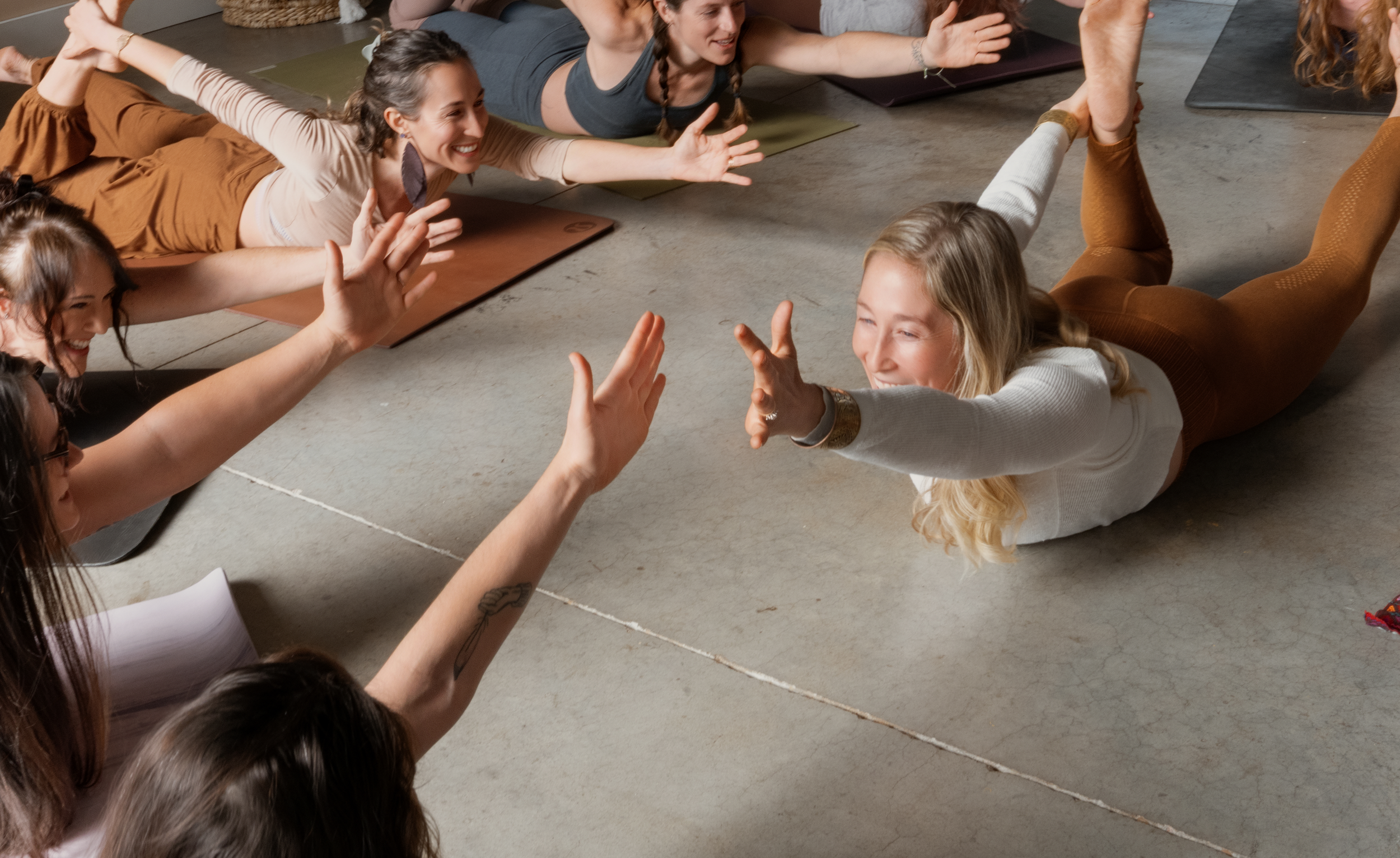 Group of women participating in a yoga or exercise class, with one woman lying on her stomach reaching out and high-fiving others, who are seated on yoga mats.