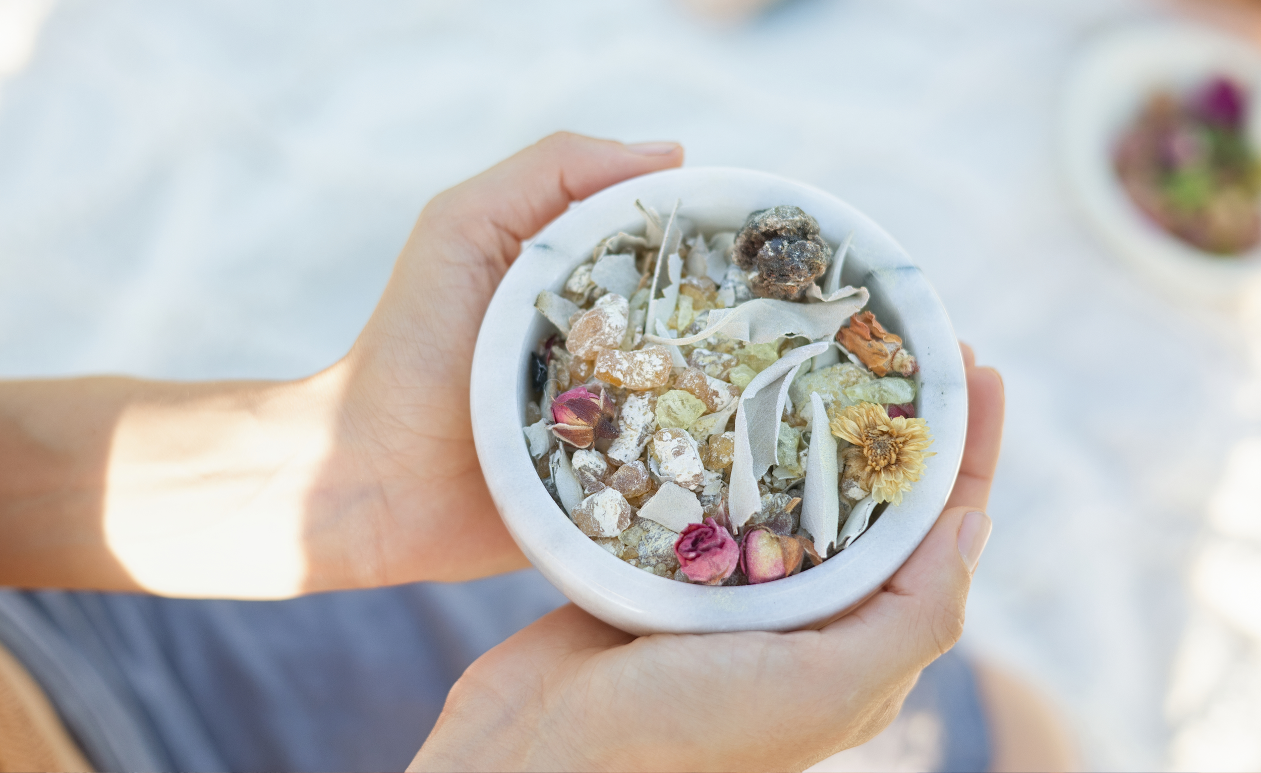 A person's hands holding a small white bowl filled with dried herbs and flowers such as rosebuds, chamomile, and lavender.