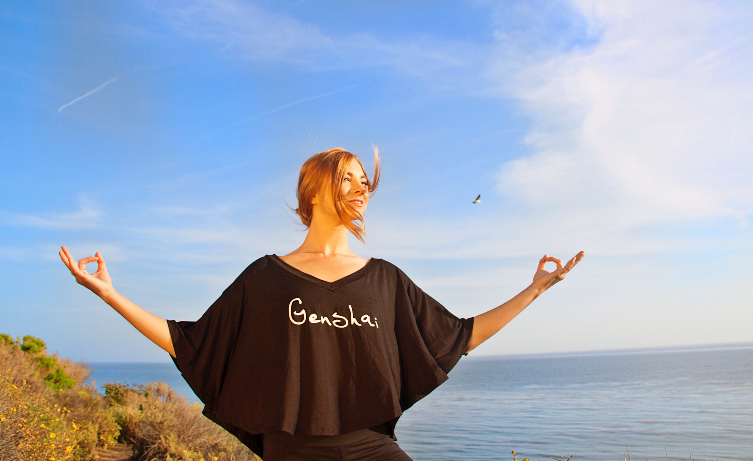 A woman standing by a body of water with a cloudy blue sky, practicing yoga or meditation with her arms raised and hands in mudra position, wearing a black top with 'Genshai' written on it.