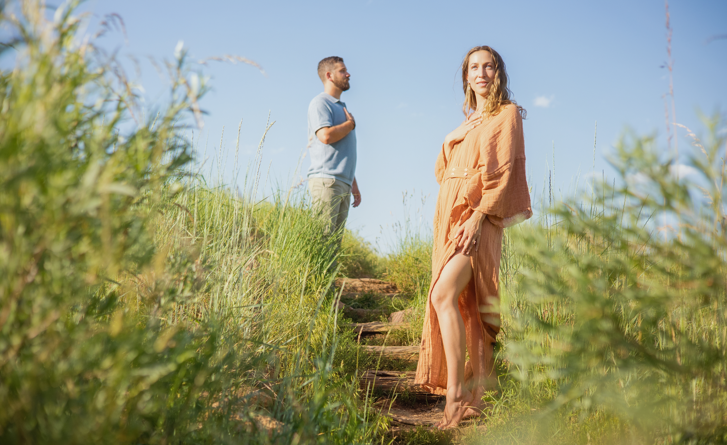 Woman in an orange dress with a slit standing barefoot on grass steps, with a man in a light blue shirt and khaki pants behind her, on a grassy hillside under a clear blue sky.