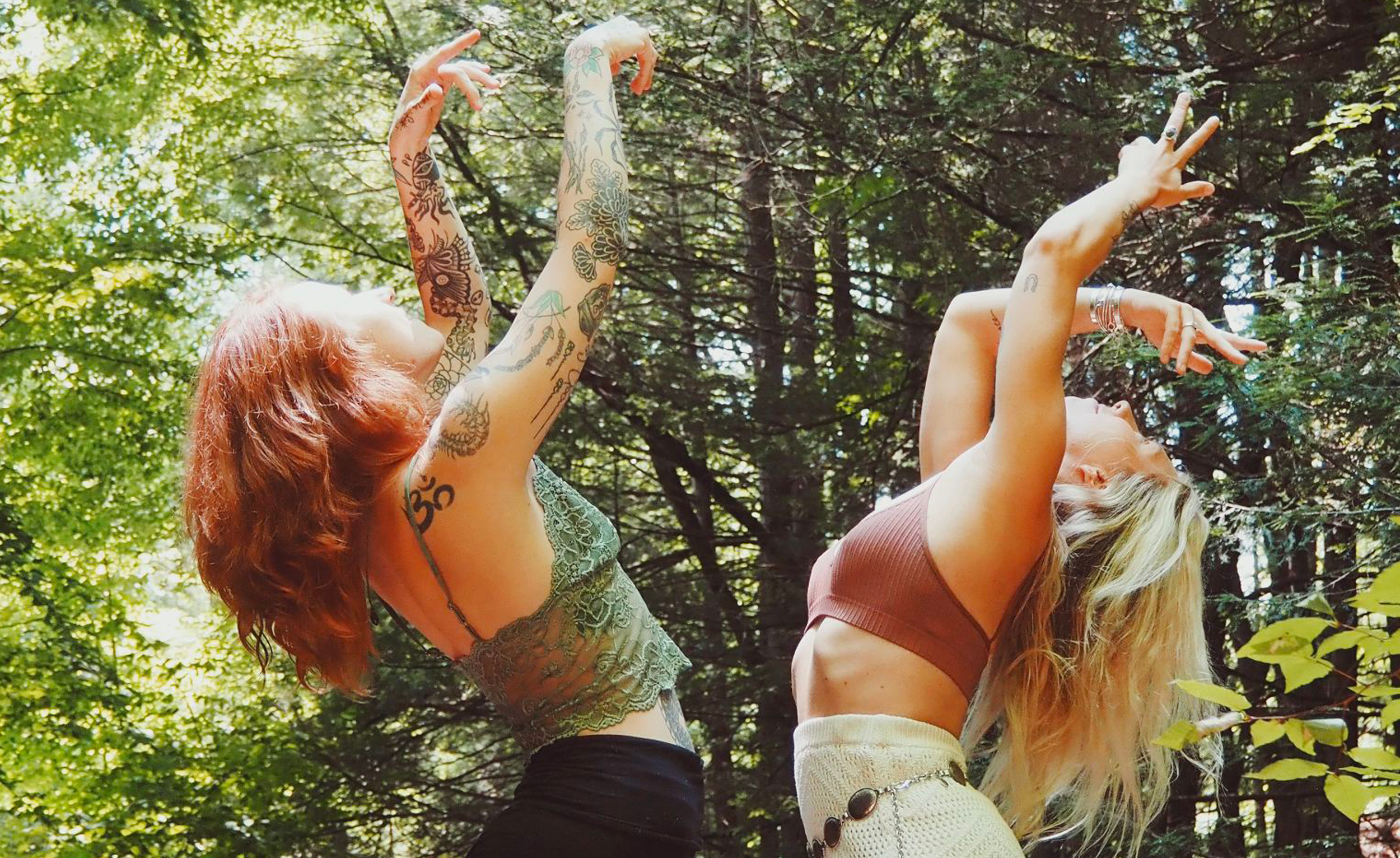 Two women dancing in a forest with arms raised and head tilted back, surrounded by green trees and sunlight filtering through the leaves.