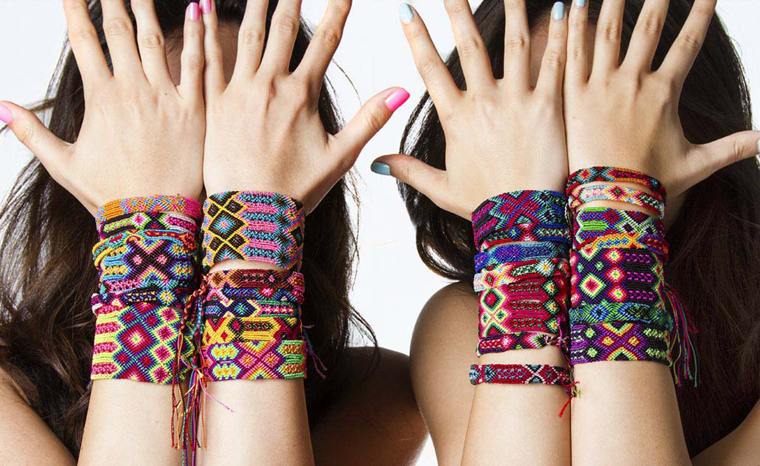 Two women with long dark hair showing colorful, patterned woven bracelets on their wrists, holding their hands up to cover their faces.