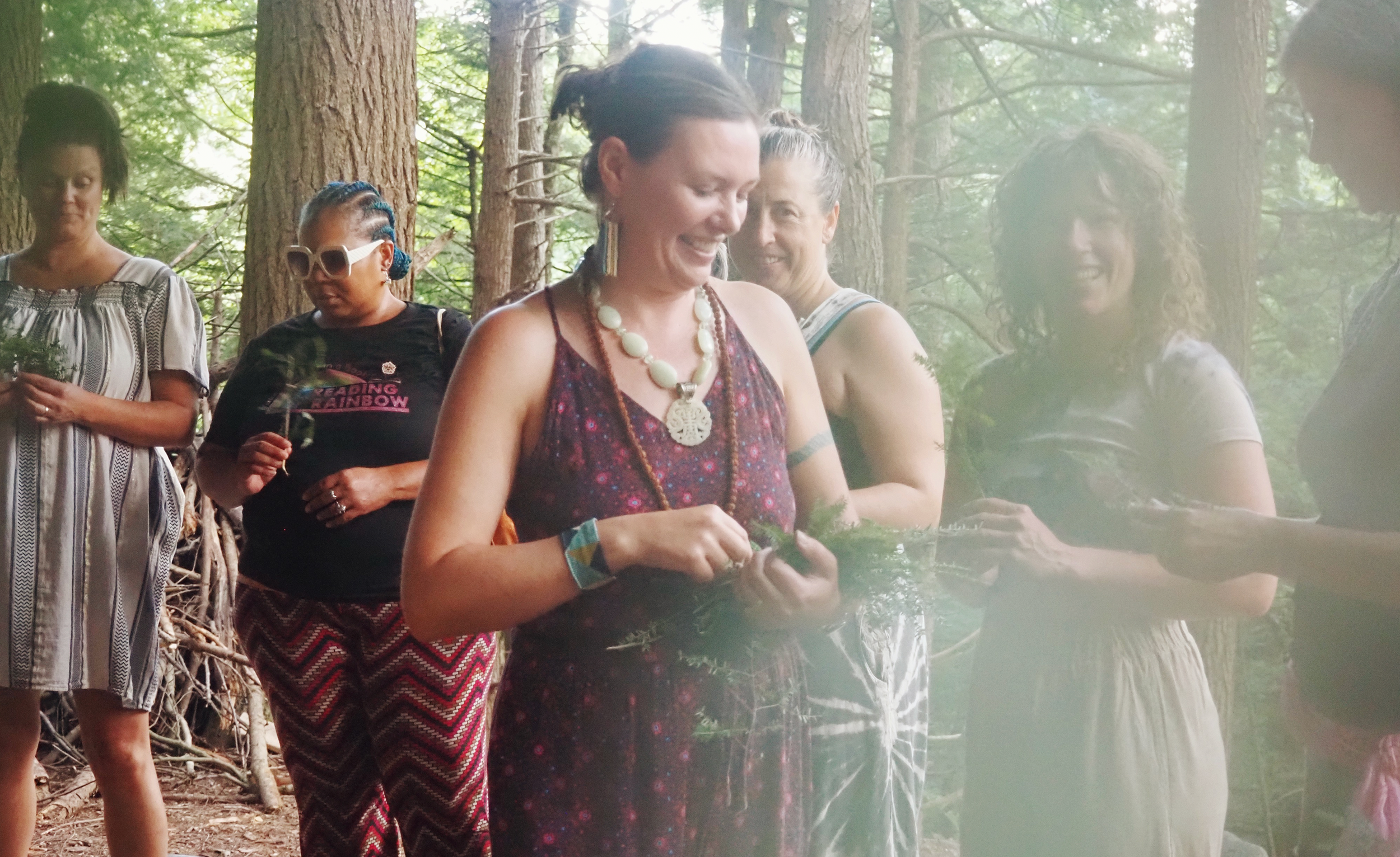 Group of women gathered in a forest, some holding greenery, engaging in an outdoor activity, smiling and interacting with each other.