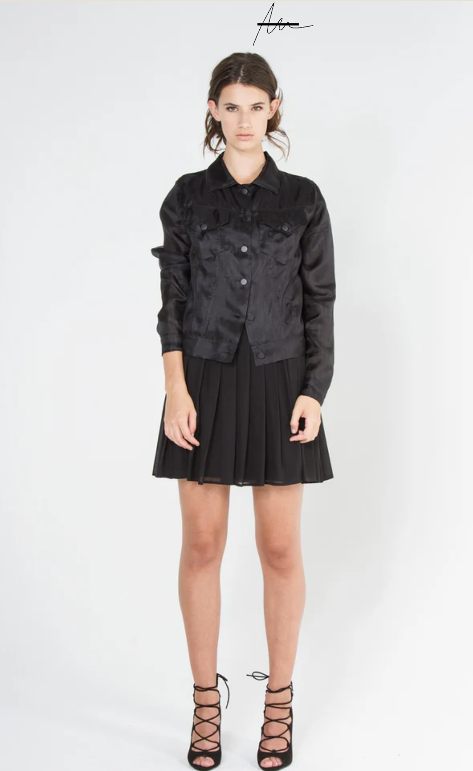 Young woman wearing a black satin jacket, black pleated skirt, and black lace-up high heels, standing against a plain white background.