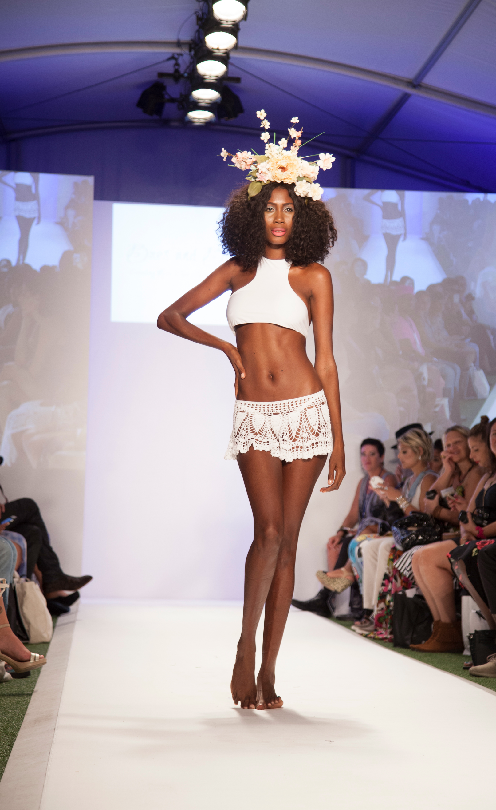 A woman with dark curly hair walks on a runway in a white crop top, white crochet shorts, and a floral headpiece, during a fashion show with an audience watching.