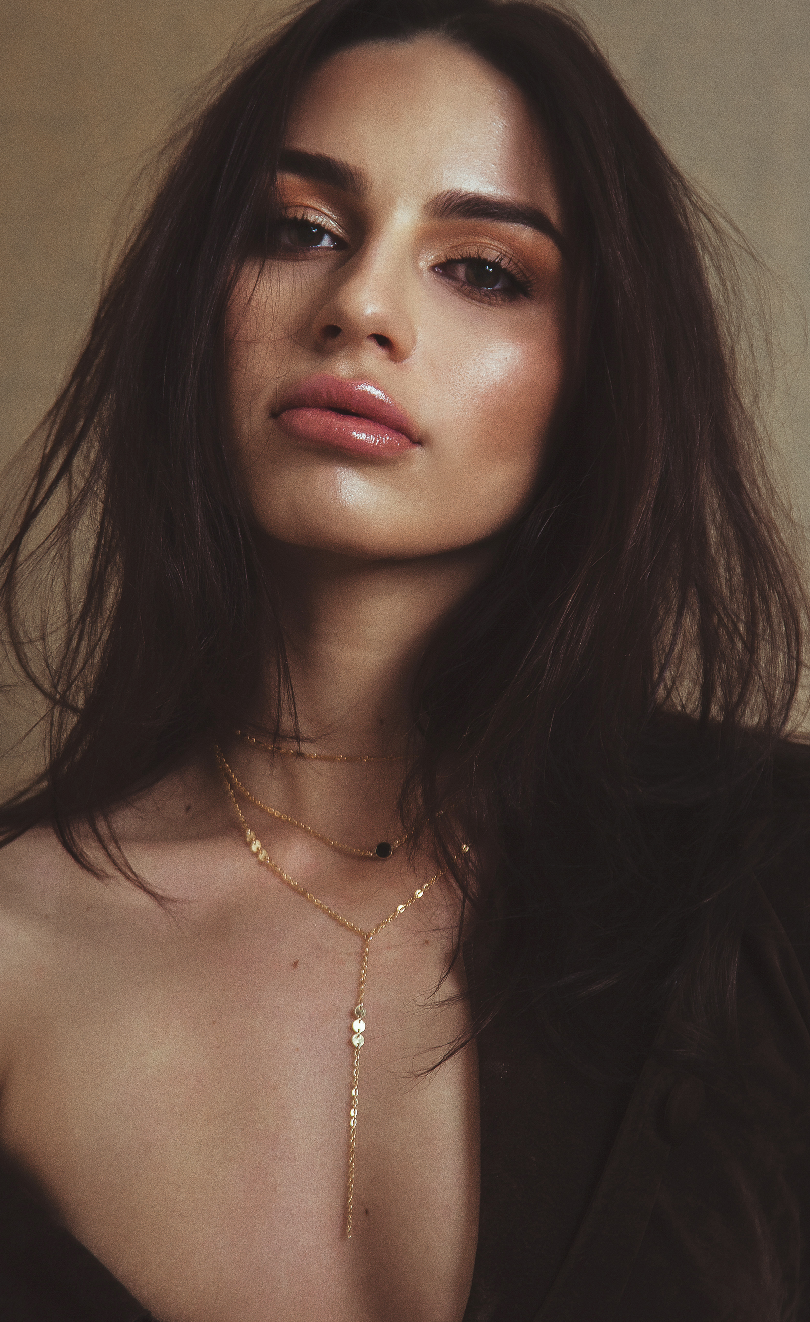 Close-up portrait of a young woman with dark hair, wearing gold layered necklaces, and a black top or dress, looking at the camera with a neutral expression.