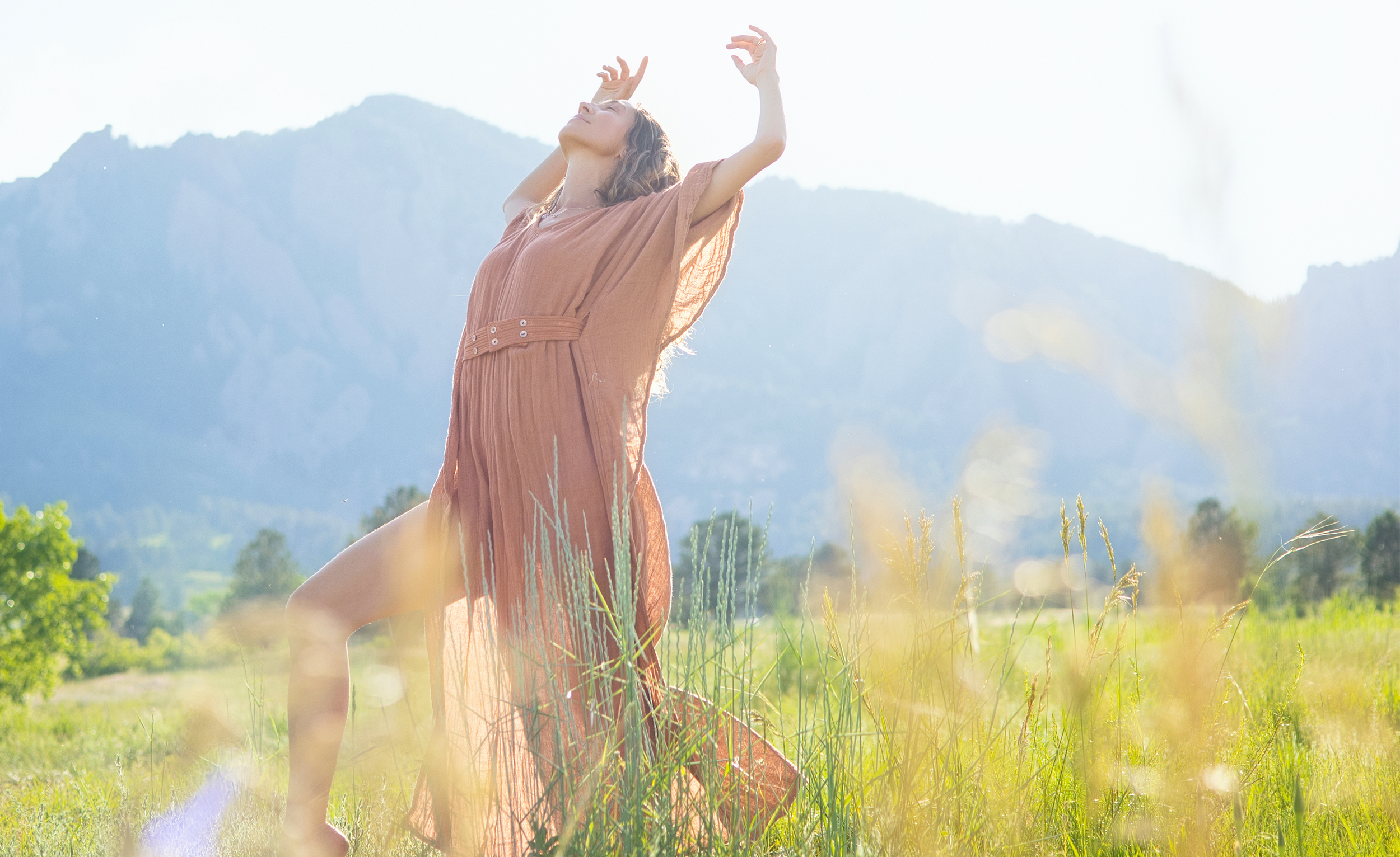 A woman wearing a flowing orange dress kneels in a grassy field with mountains in the background, sunlit, raising her arms in an expressive posture.