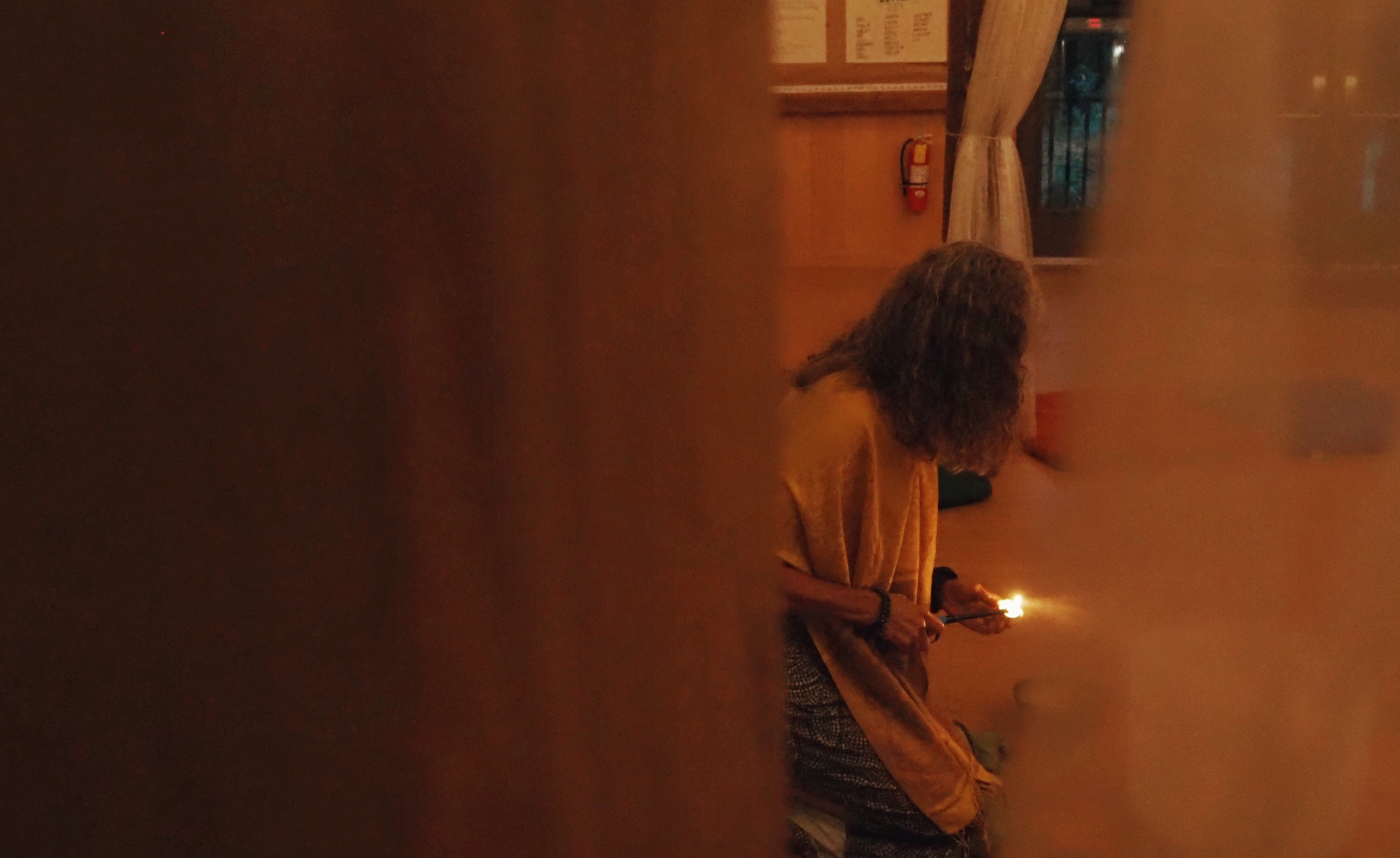 A person with curly hair wearing traditional clothing, sitting indoors, holding a lit match or sparkler, illuminated by its glow, with a partially visible person in the foreground and a wooden interior background.