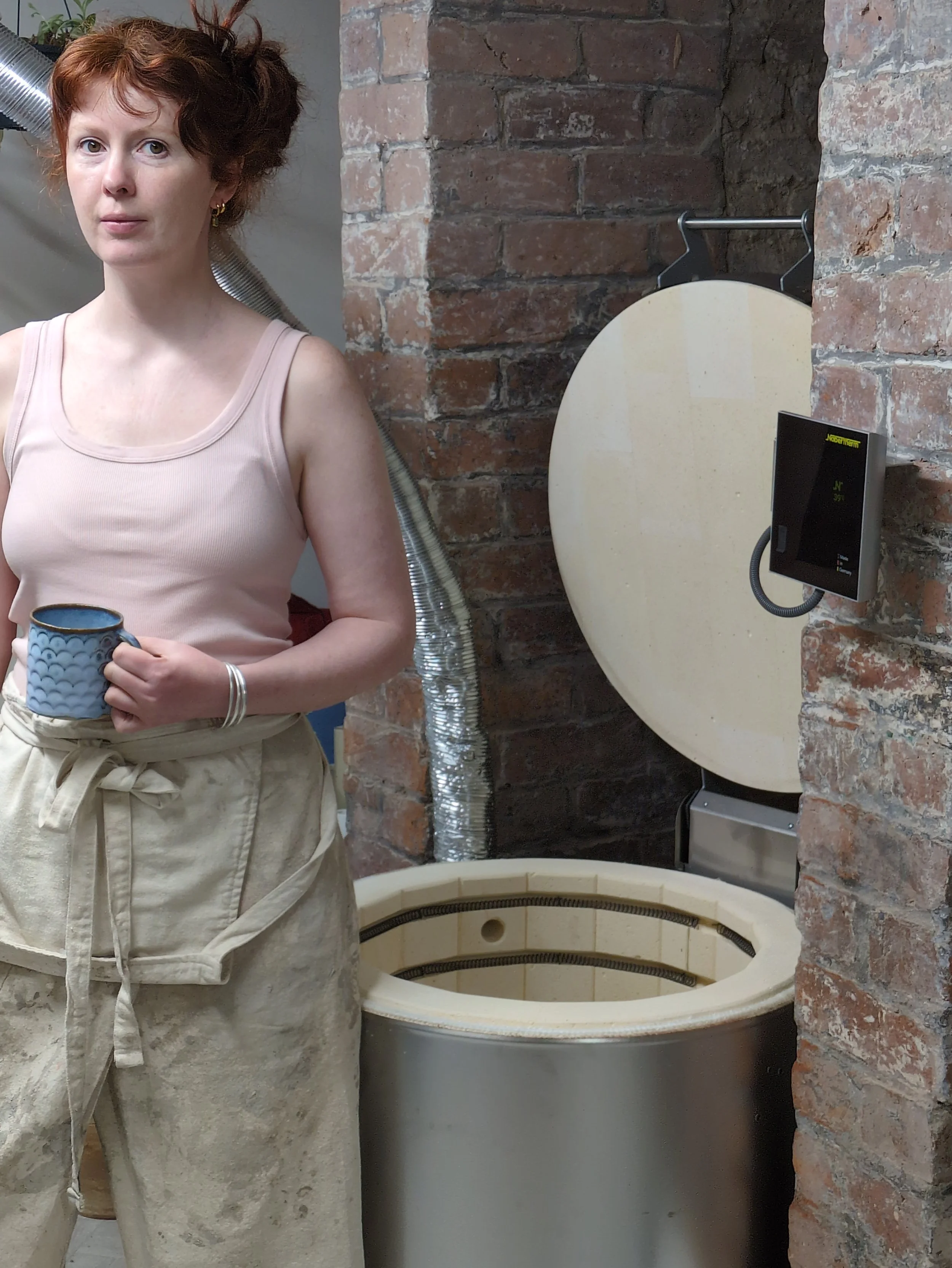 Ceramic artist in pottery workshop holding a handmade mug next to a kiln in studio.