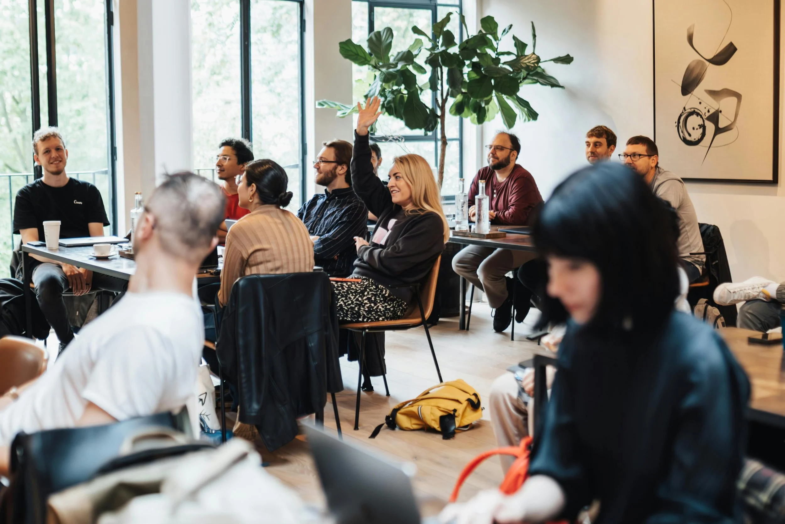 A diverse group of people attending a meeting or workshop in a bright room with large windows, some taking notes, one woman raising her hand.