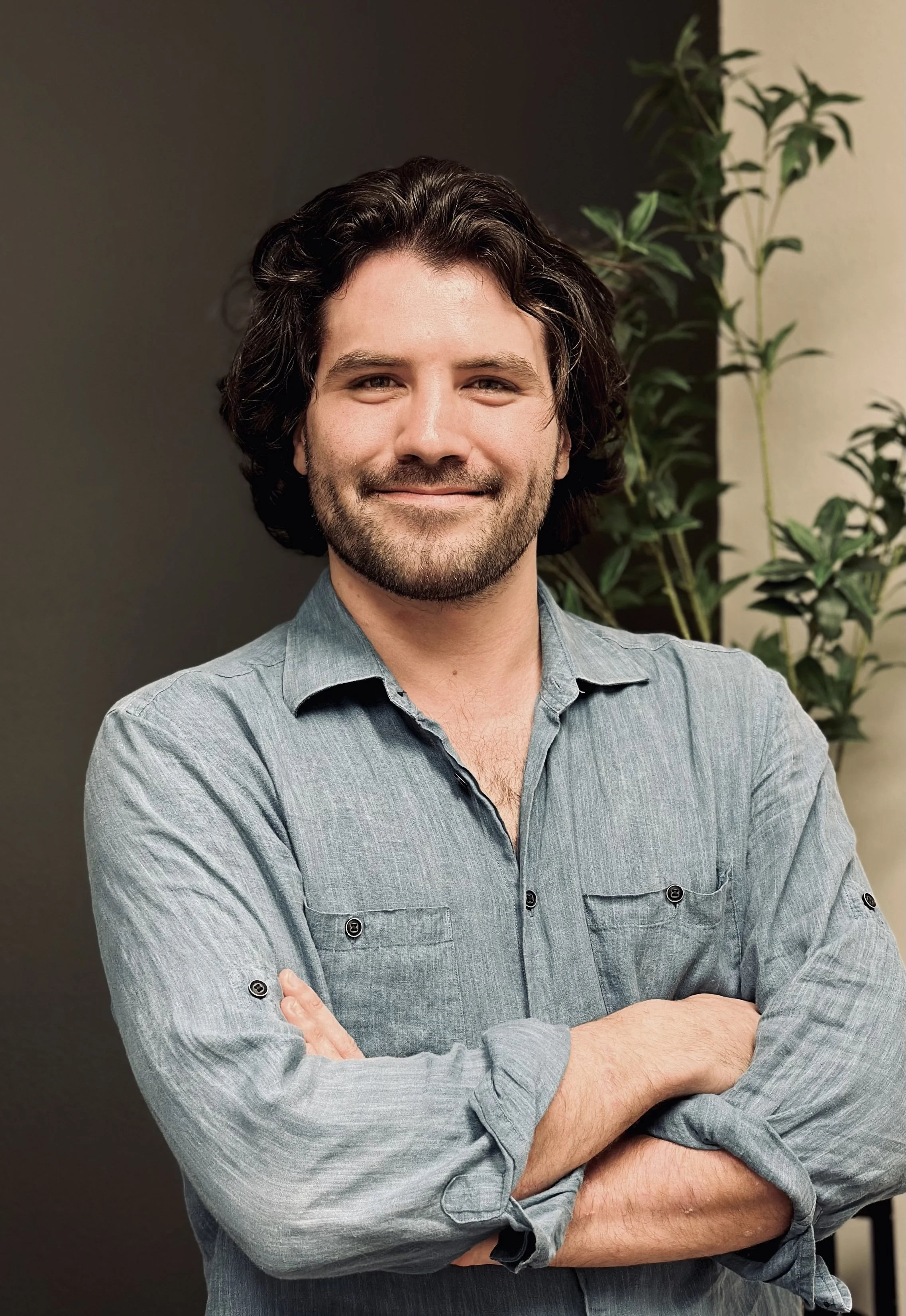 A smiling man with dark, wavy hair and a beard, wearing a light gray button-up shirt with rolled-up sleeves, standing in front of a dark wall and a leafy green plant.