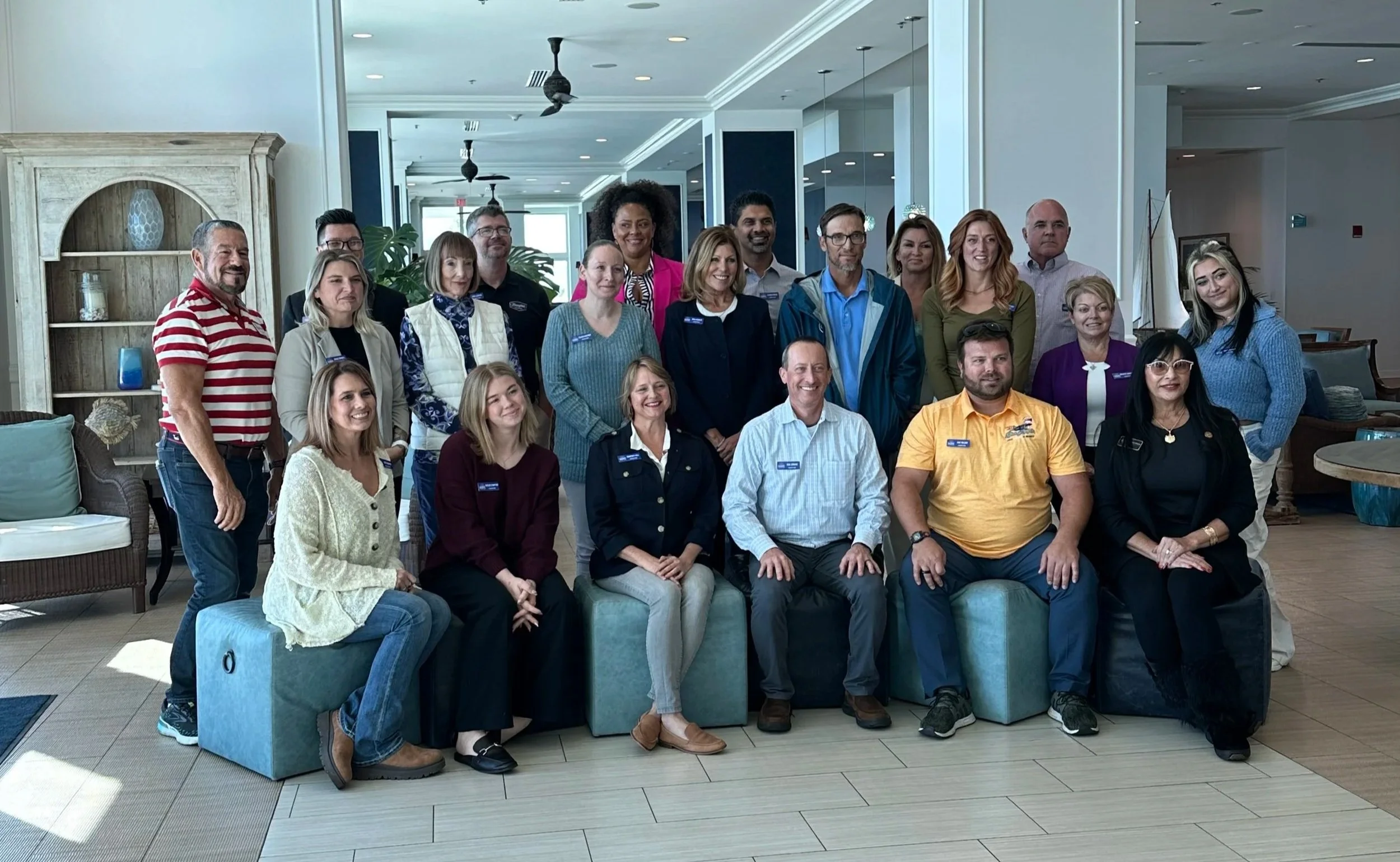 A group of approximately 20 people, diverse in age, gender, and ethnicity, posing together indoors in a well-lit, modern space with a large mirror, wooden furniture, and decorative items in the background.