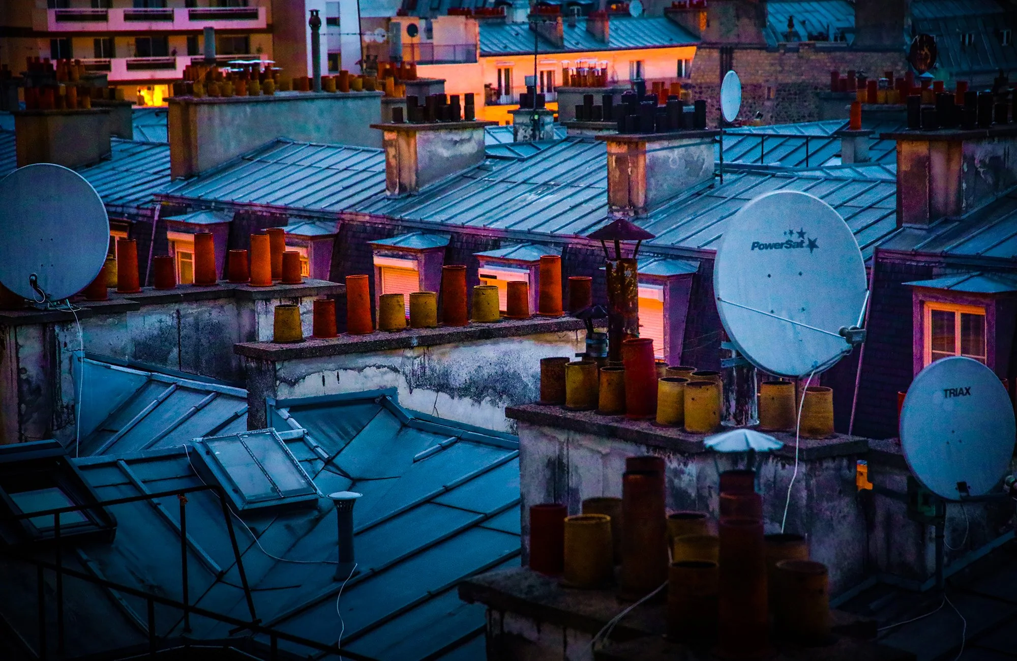 Rooftops of city buildings with satellite dishes, chimney pipes, and skylights during twilight.