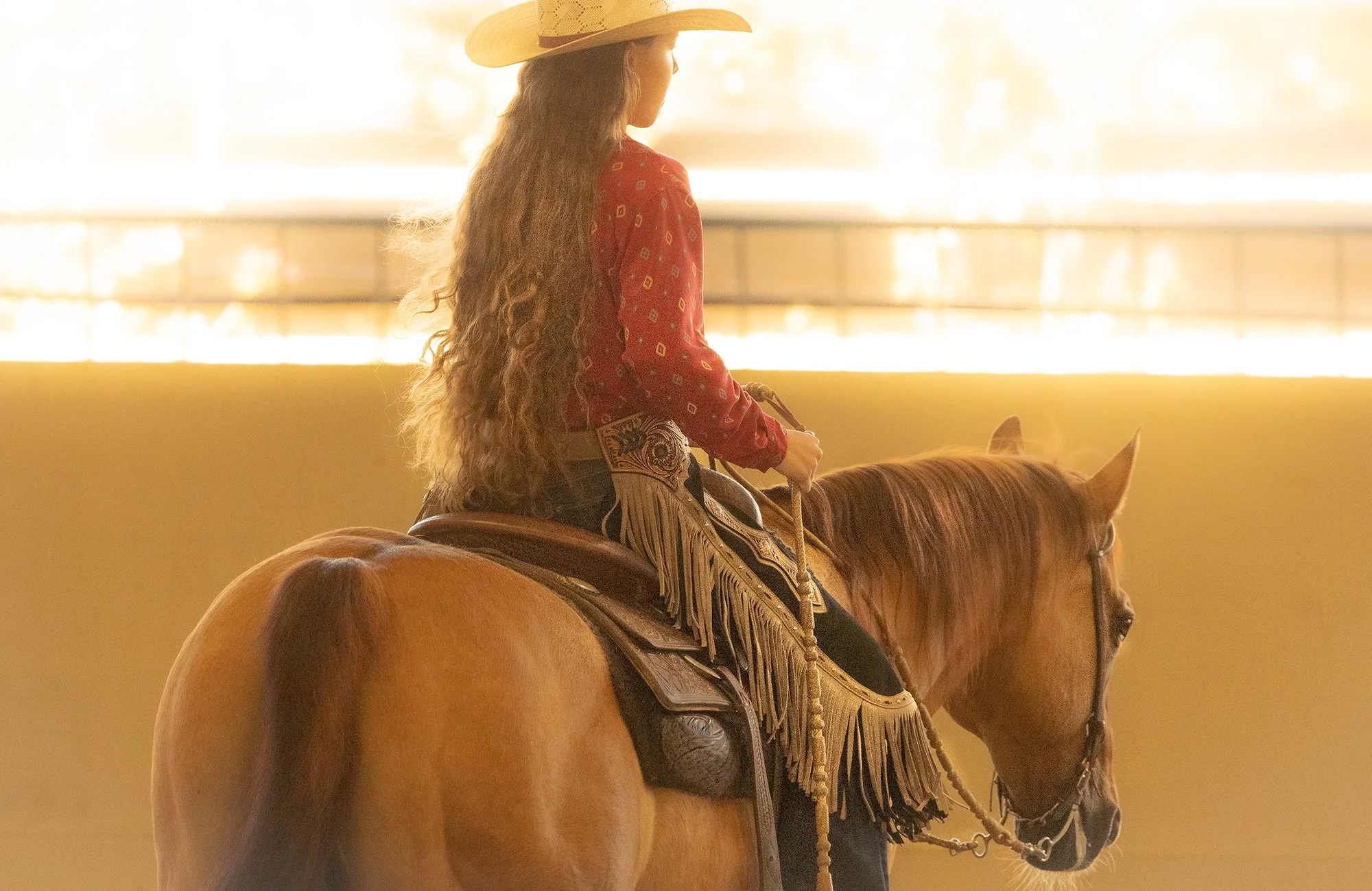 This is a warm, beautifully backlit photograph of a cowgirl riding a chestnut horse, likely inside an indoor arena or under a covered pen. The image is captured from behind and slightly to the side, highlighting the rider's long, wavy hair cascading 