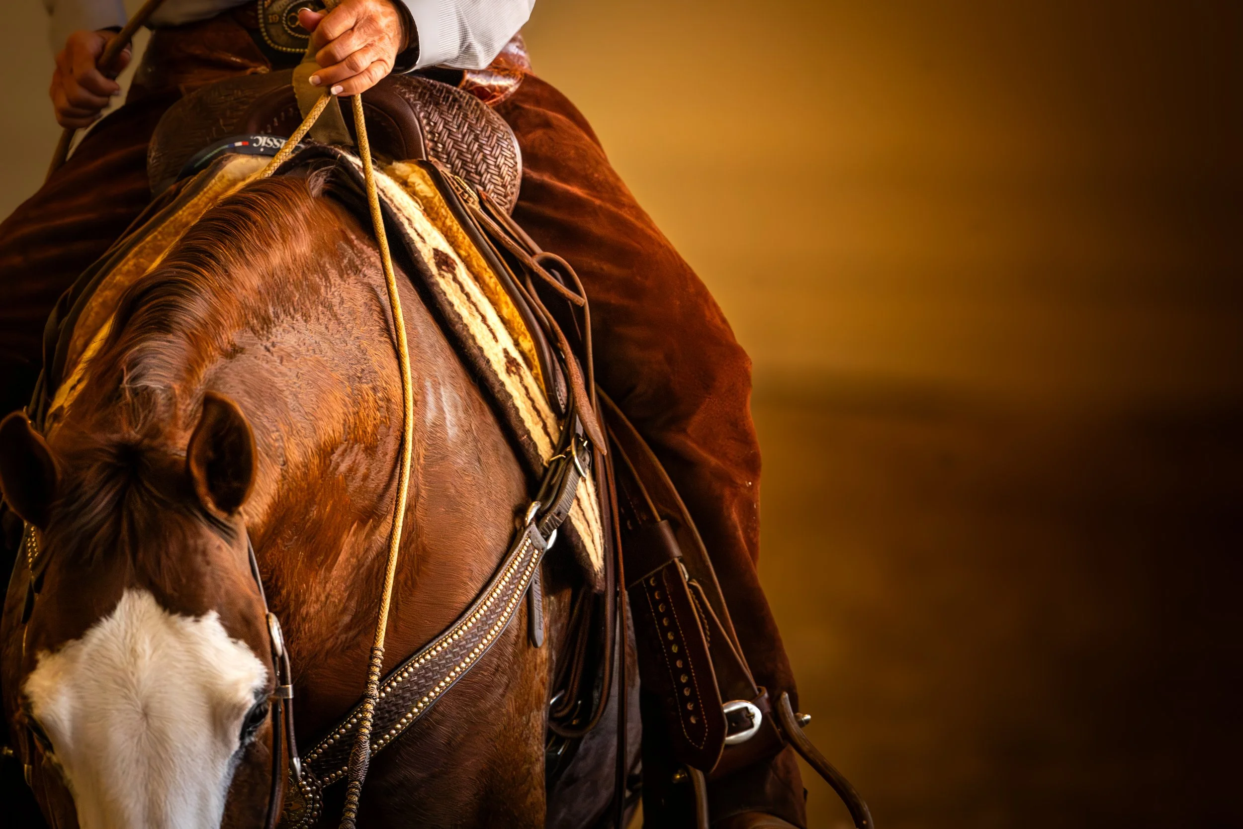 This is a rich, warm-toned close-up photograph focusing on the intricate details of Western riding. The image captures the torso and legs of a rider seated on a chestnut horse with a prominent white blaze on its face. The rider is wearing a light-col