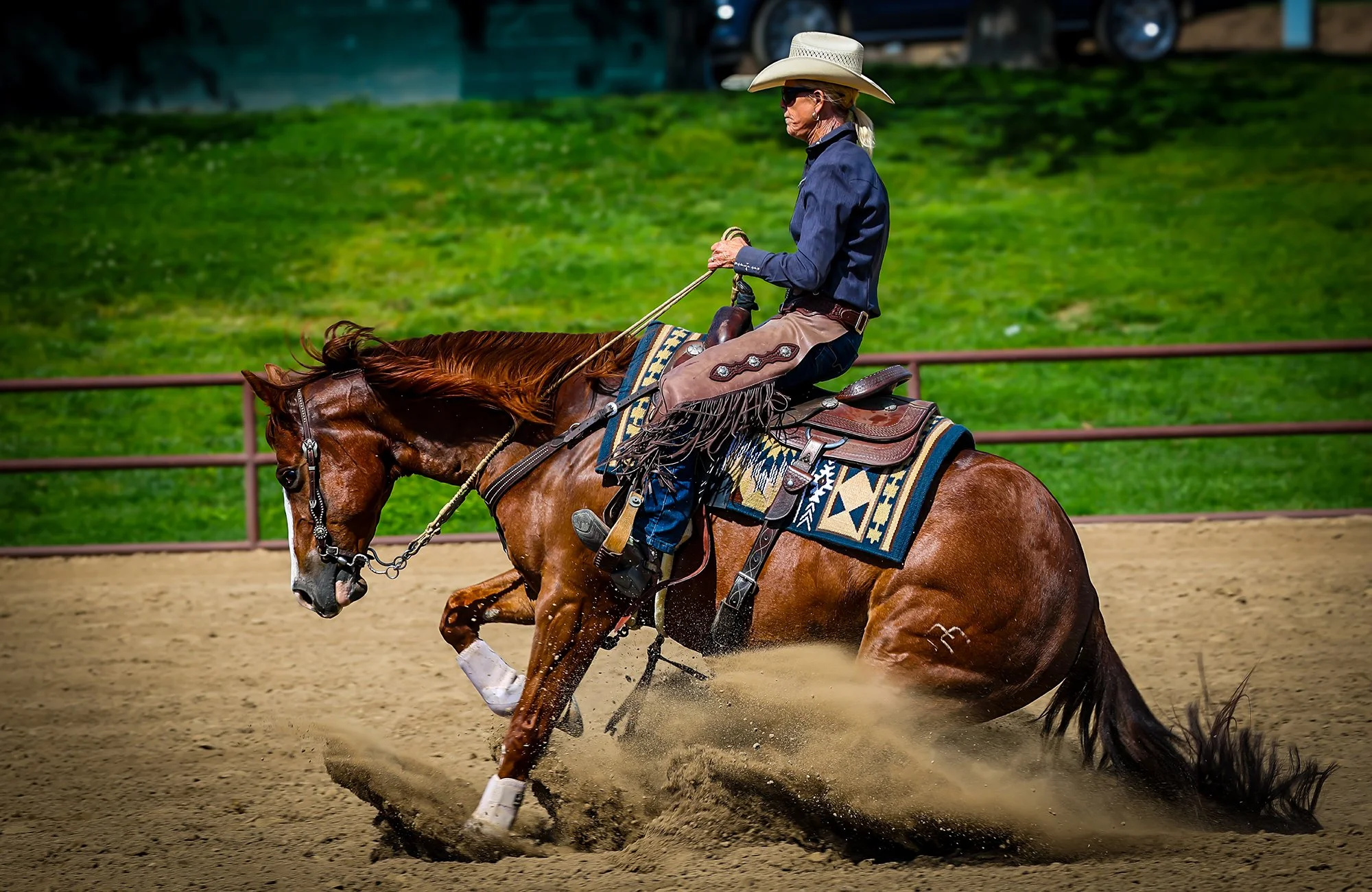 This is an action-packed image of a female equestrian riding a beautiful chestnut horse in an outdoor arena. The horse is in the middle of a dynamic "sliding stop," a classic maneuver in Western reining, kicking up a dramatic cloud of dirt from its h