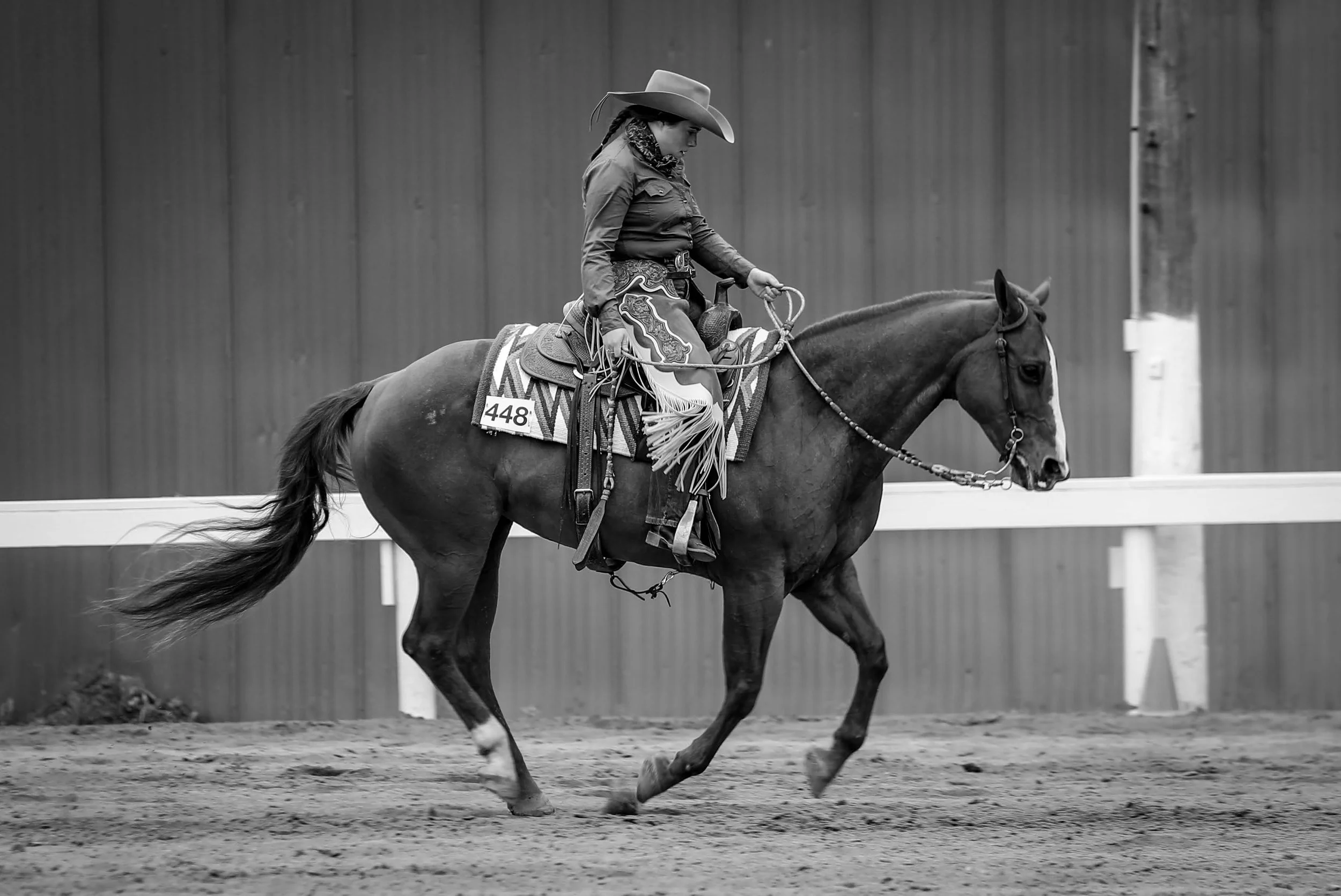 A woman in cowboy attire riding a horse in an arena, with a wooden wall in the background.