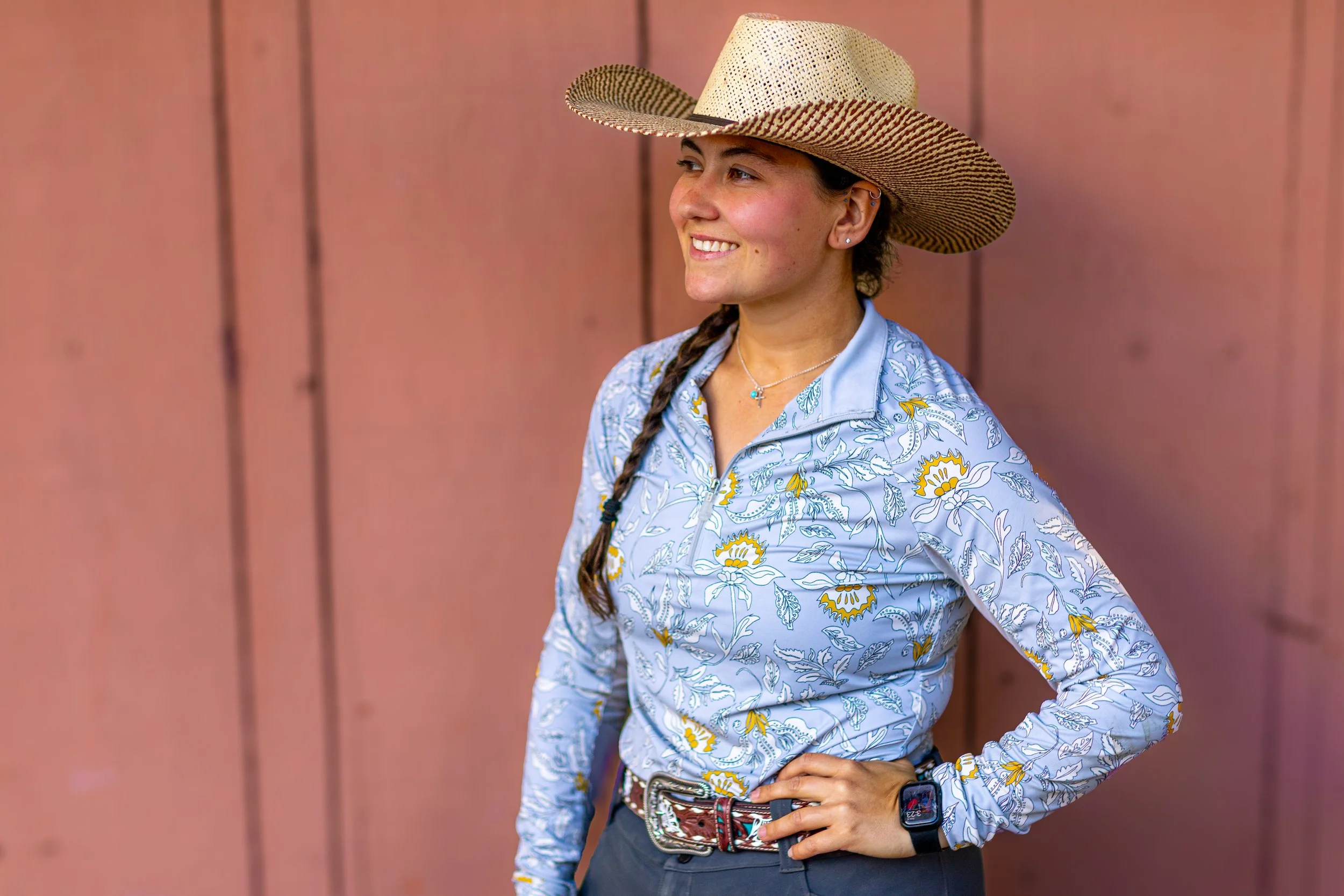 A woman wearing a straw wide-brimmed hat, a floral longsleeve shirt, a belt, and a smartwatch, standing against a pink wall.