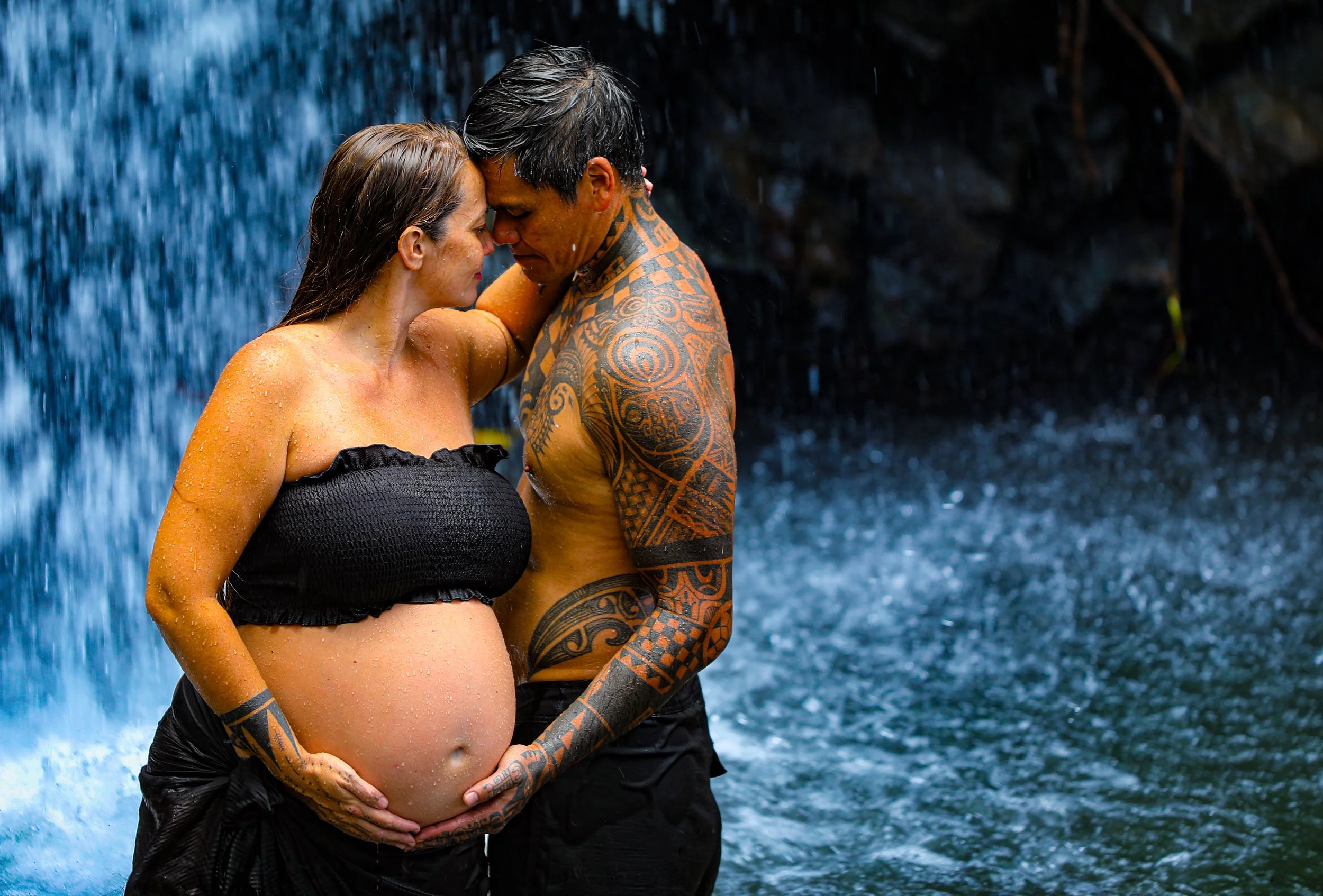 A pregnant woman and a tattooed man sharing an intimate moment under a waterfall, with their foreheads touching and hands on her belly.