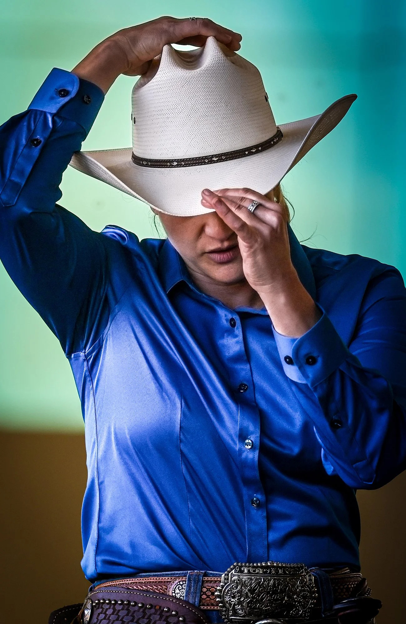 This captivating close-up portrait features a Western equestrian, seen from the chest up, in a moment of candid action or contemplation. The person is wearing a crisp white straw cowboy hat, which they are either adjusting or using to shade their eye
